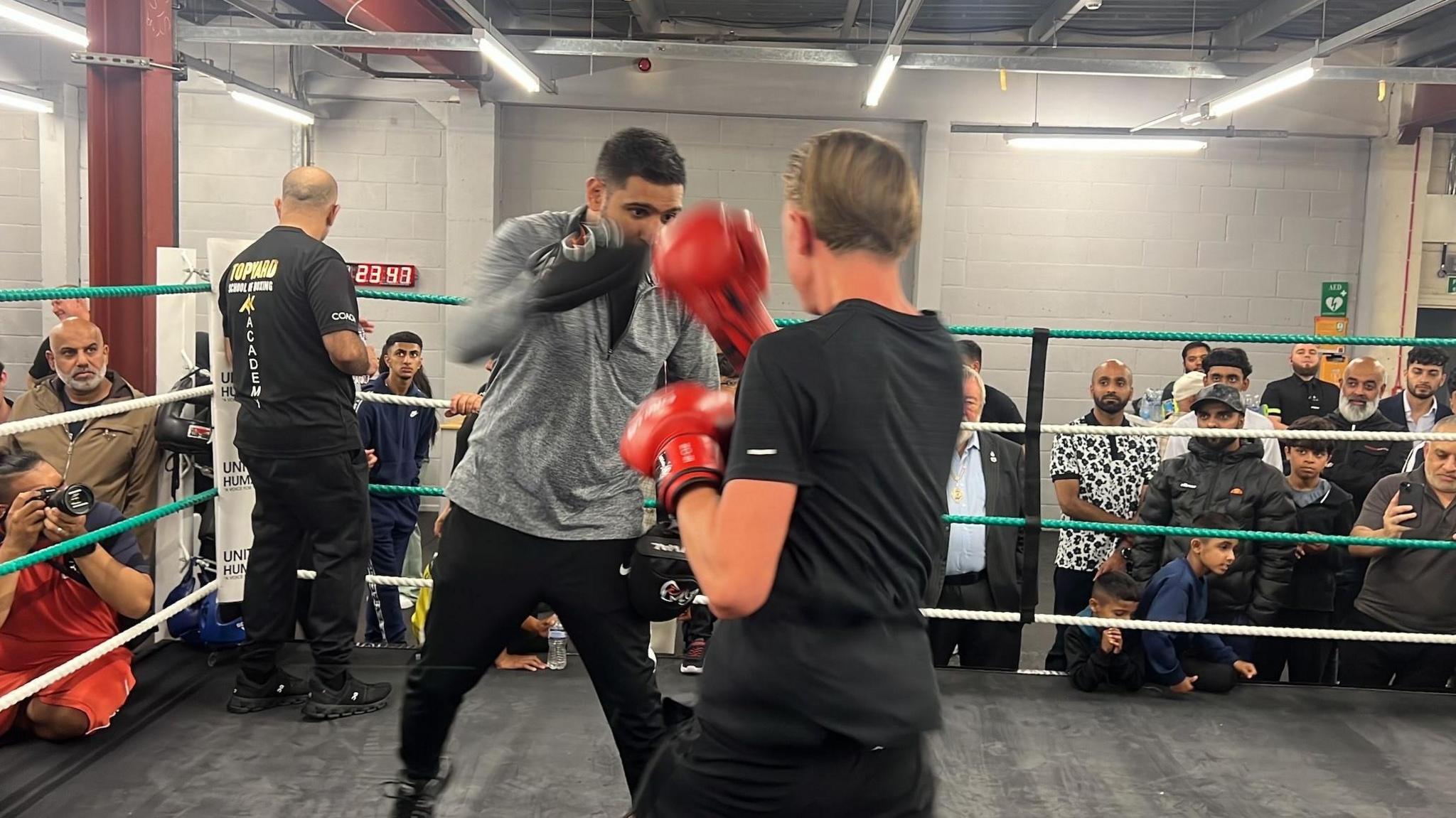 Khan, who is wearing a grey T-shirt and black trousers, is holding his hands up which have boxing gloves on. A person is standing in front of him with their back to the camera wearing red boxing gloves.