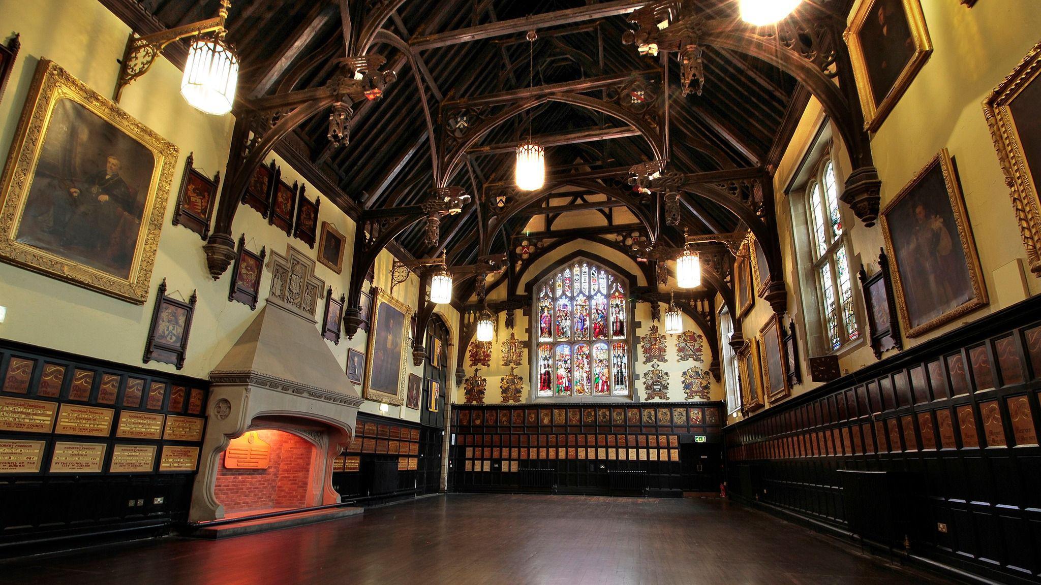 The Great Hall is a grand historic room. A large arched stain glass window sits on one side of the room. There is a large stone fireplace and the walls are lined with dark wood designs.