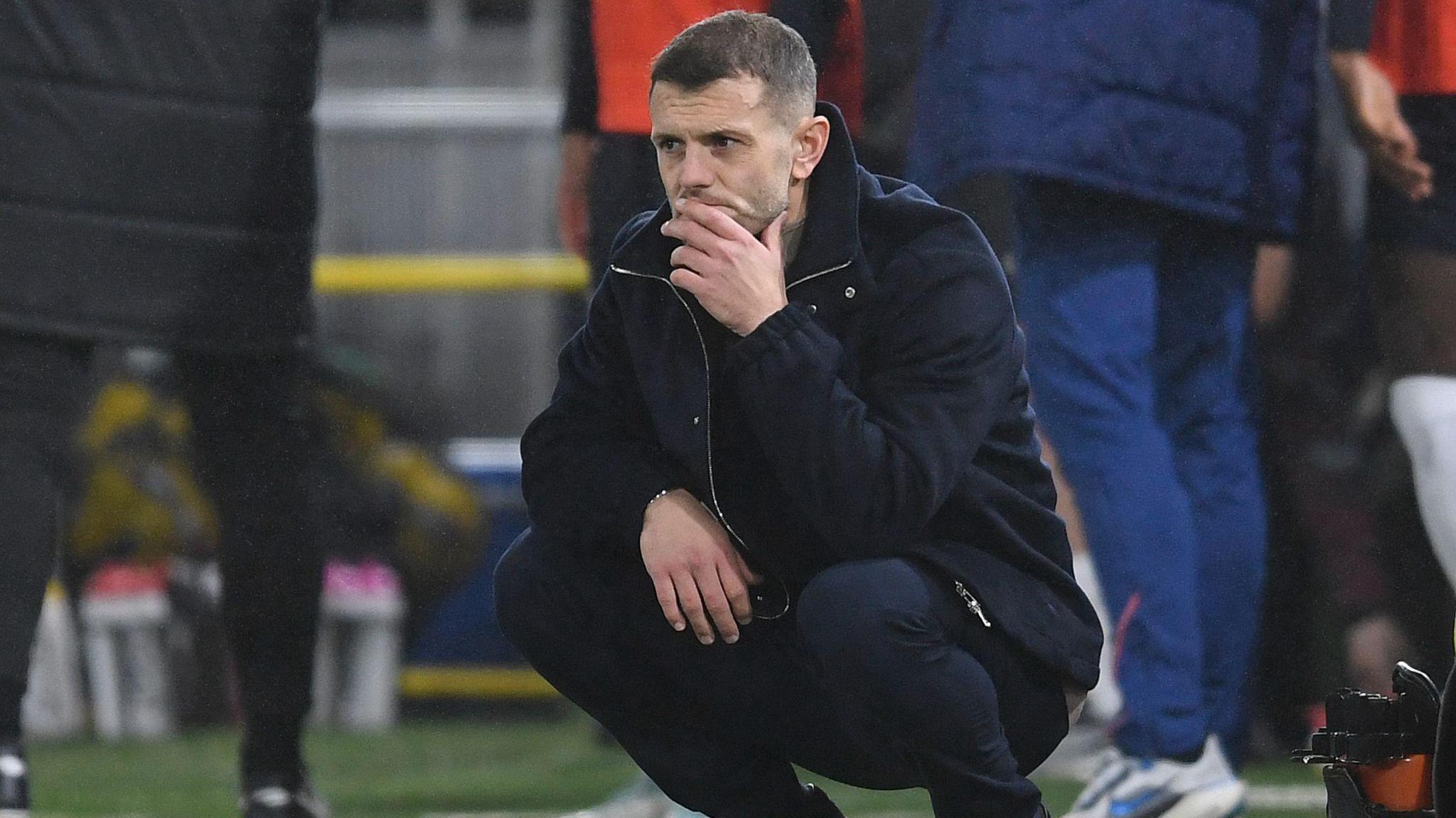 Jack Wilshere crouches down and strokes his chin in the technical area