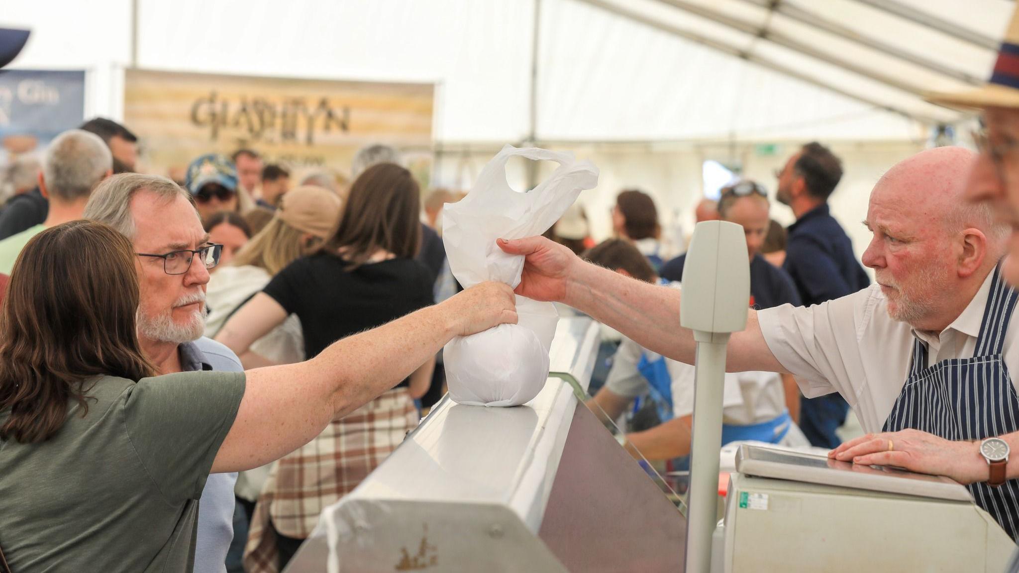A butcher passes a plastic bag to a woman over a counter in a crowded white marquis.