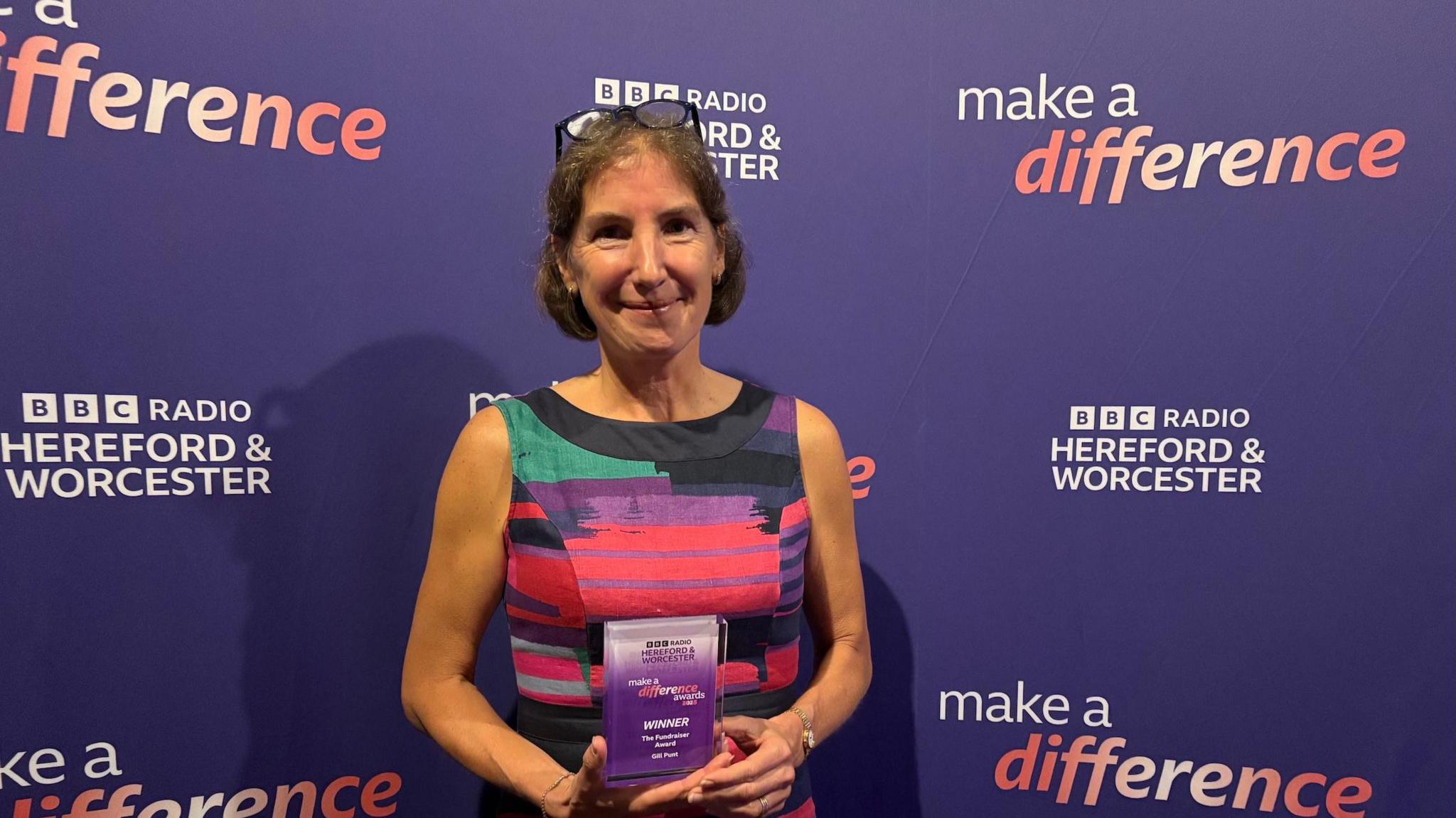 A woman with short brown hair wearing a multicoloured sleeveless dress. She has black rimmed glasses sitting on her head and is holding a small rectangular trophy. She is stood in front of a board that reads BBC Radio Hereford and Worcester and make a difference.