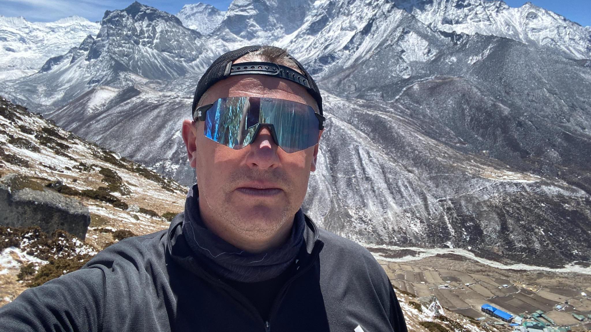 A selfie of Mark Rowlands which shows him standing in the Himalayas. There are snow-capped mountains behind him and he is wearing blue reflective ski glasses and a dark-coloured fleece. He is also wearing a black cap which is on back-to-front.