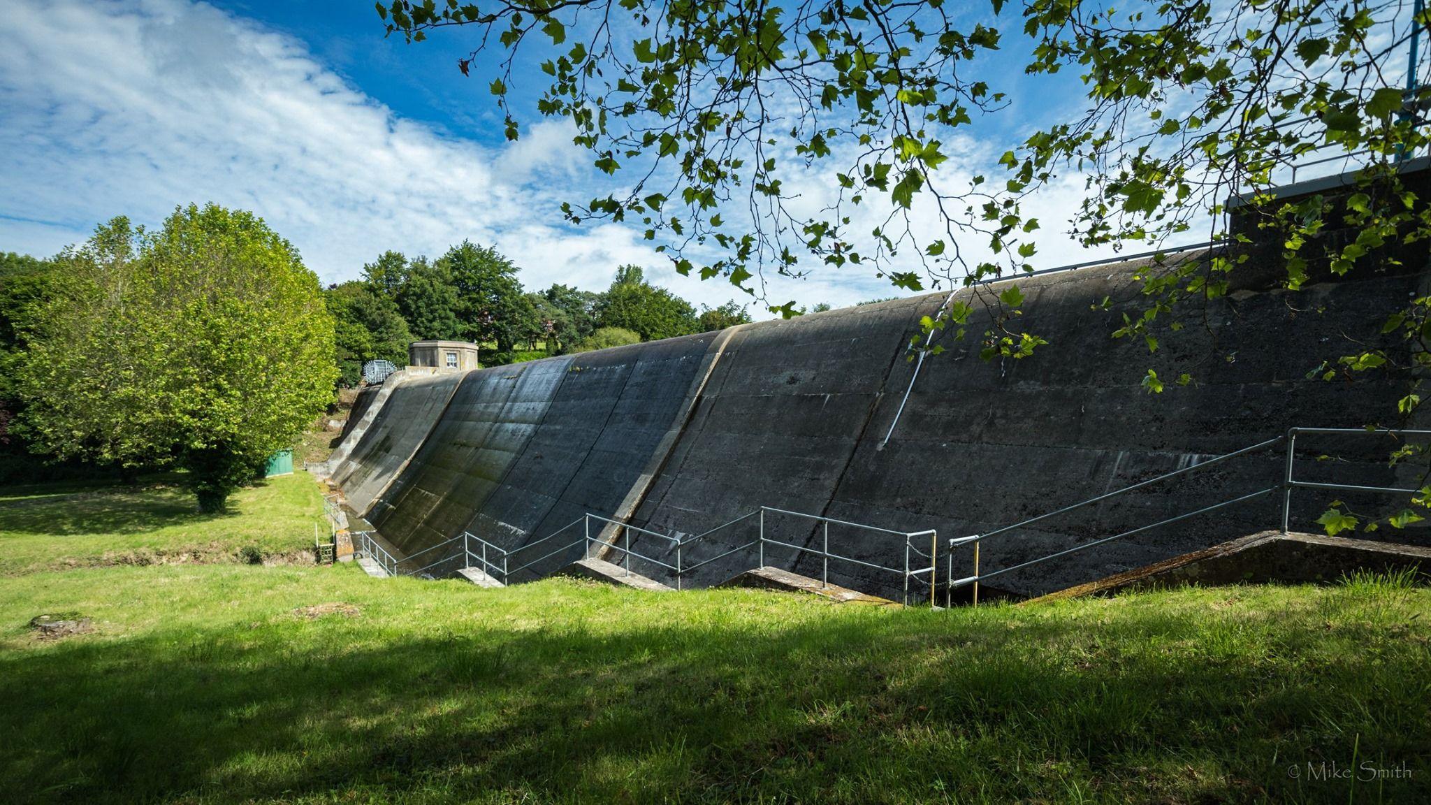 The image depicts a large concrete dam with a sloped face, surrounded by green grass and trees. Metal railings and stairs run along the side of the dam, leading down to its base. A small building or control room is visible on top of the dam, partially hidden by trees. The sky is mostly clear with a few scattered clouds, and some tree branches are visible in the foreground.