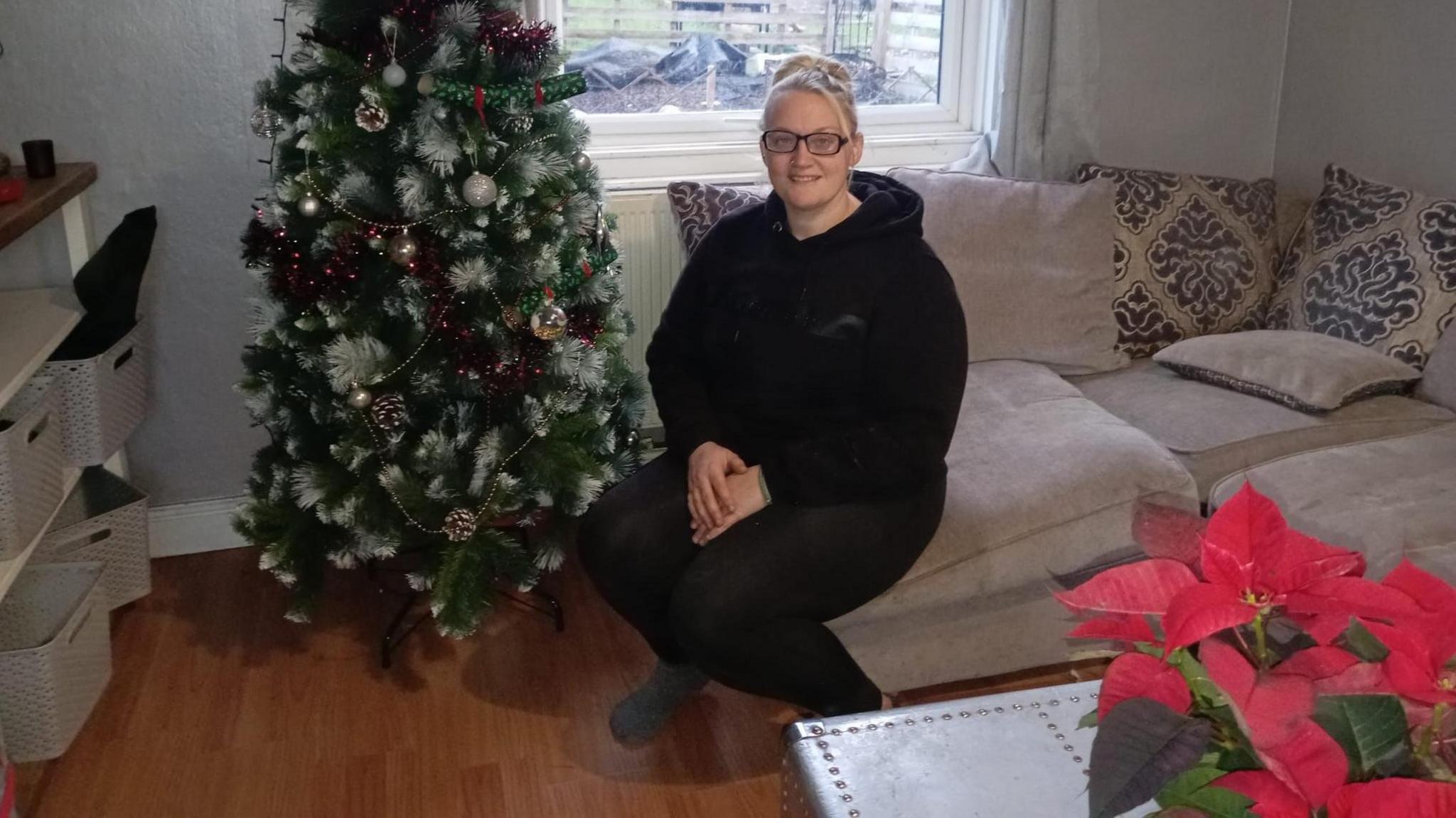 Sidony West, a blonde woman with glasses, sits in her living room on a sofa next to a Christmas tree. 