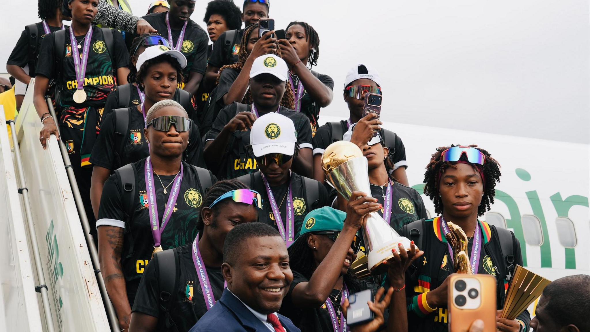 Cameroon players, predominantly wearing black t-shirts and gold medals on purple ribbons around their necks, stand on the steps of an airplane as the Women's Africa Cup of Nations trophy is held aloft. The head of a smiling man in a suit is seen in front of the players at the bottom of the stairs