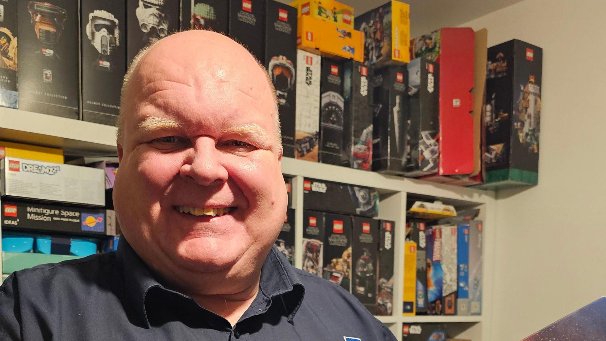 A man in a black shirt holds a Lego kit standing in front of shelves with many Lego kits on. 