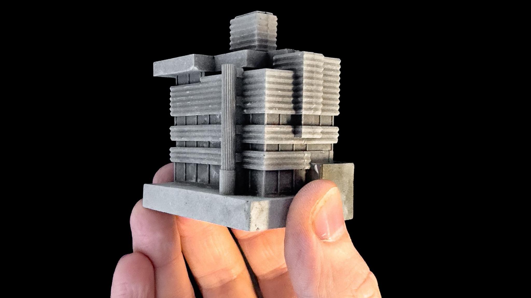 A model of the New Street signal box which is held by a hand in front of a black background. The model shows various windows and curved surfaces representing the building in grey concrete.