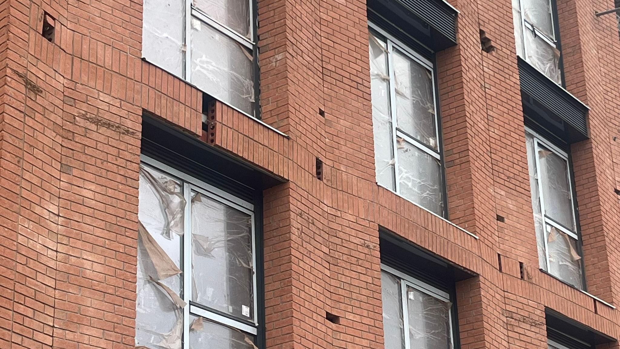 A closer view of the building with bricks missing from the front of the building with plastic wrap on the front of the windows.