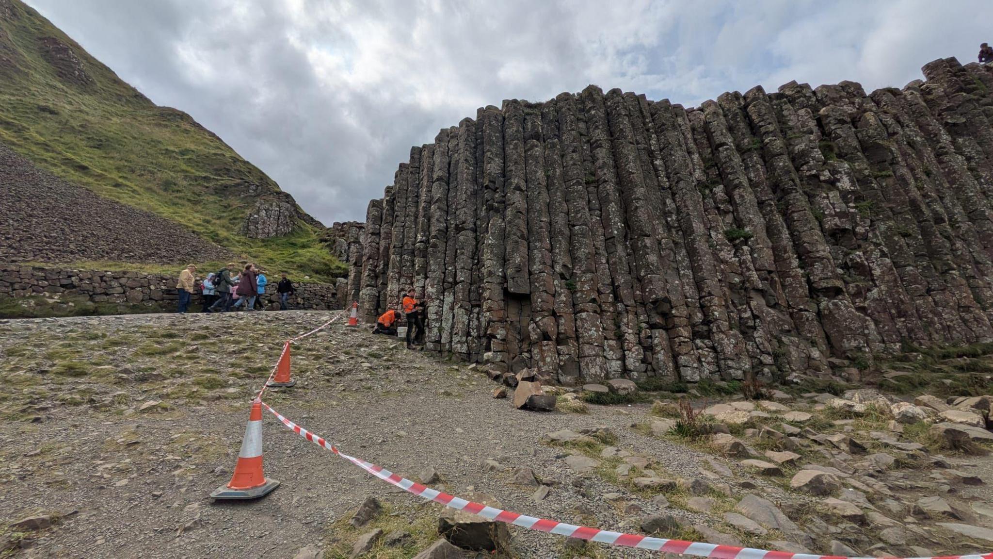 Tape and cones are cordoning off an area beside tall basalt columns. Some people are standing nearby. Some workers in high vis jackets are beside the rocks.