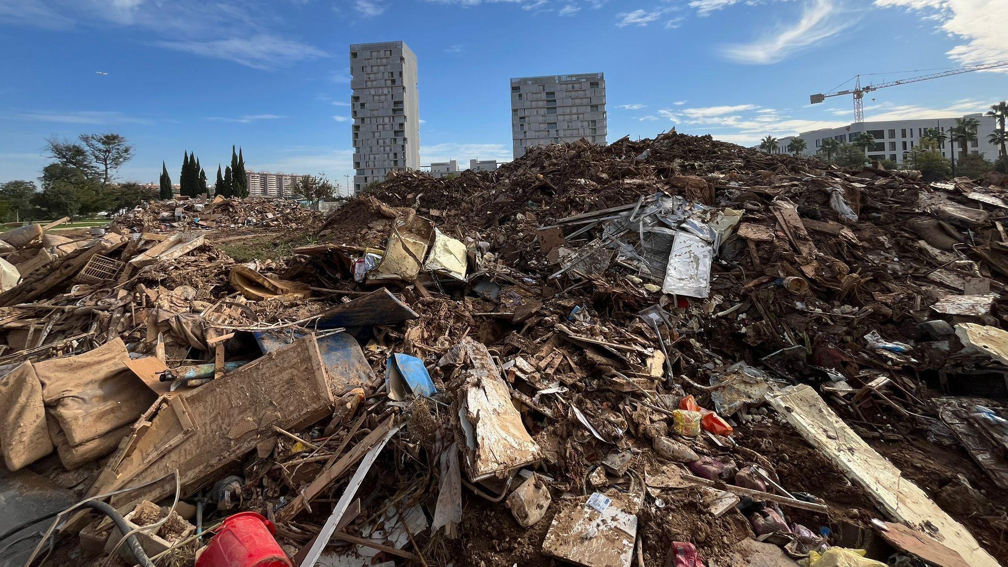 A massive pile of dirt, furniture and debris that has been swept up and deposited following the flash floods in Valencia. In the background there are three tall buildings and some trees still standing, but everything else around them has been destroyed.