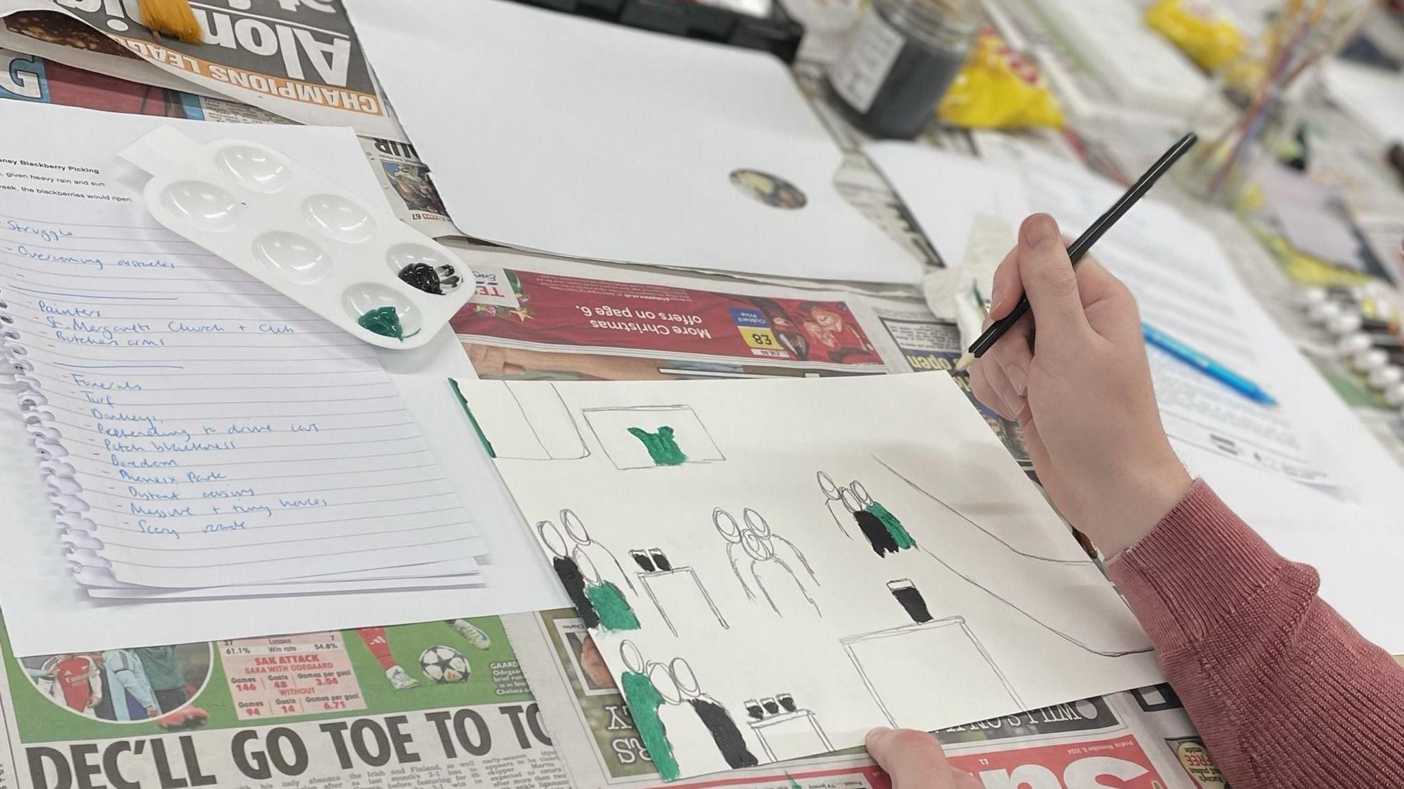 A table covered in newspapers and the arm of someone using a pencil and paintbrush to design a picture showing groups of people in a pub. The people are wearing green and black clothes. There is a paint pallet with green and black paint, and a piece of notepaper with a bullet point list