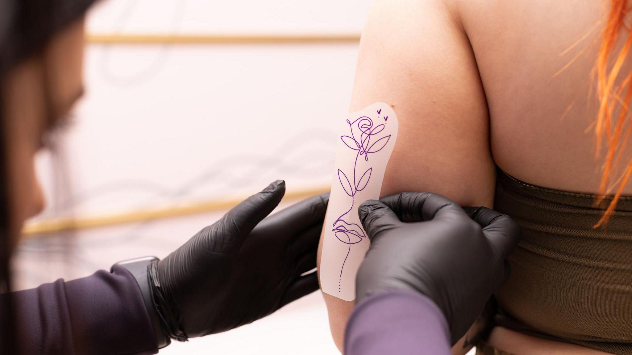 A female tattoo artist wearing black latex gloves and laying a minimalist line drawing stencil on the back of a woman's arm prior to tattooing