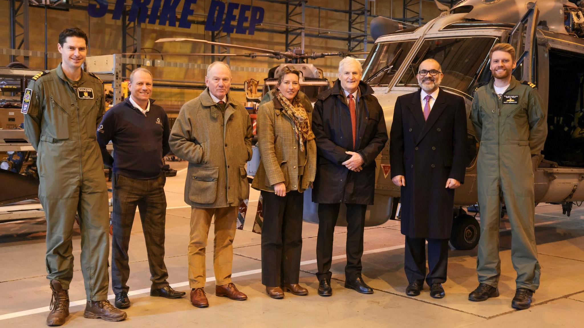 Seven people are standing in a row in front of a navy helicopter. Two of the people are in navy unform, some are wearing suits.