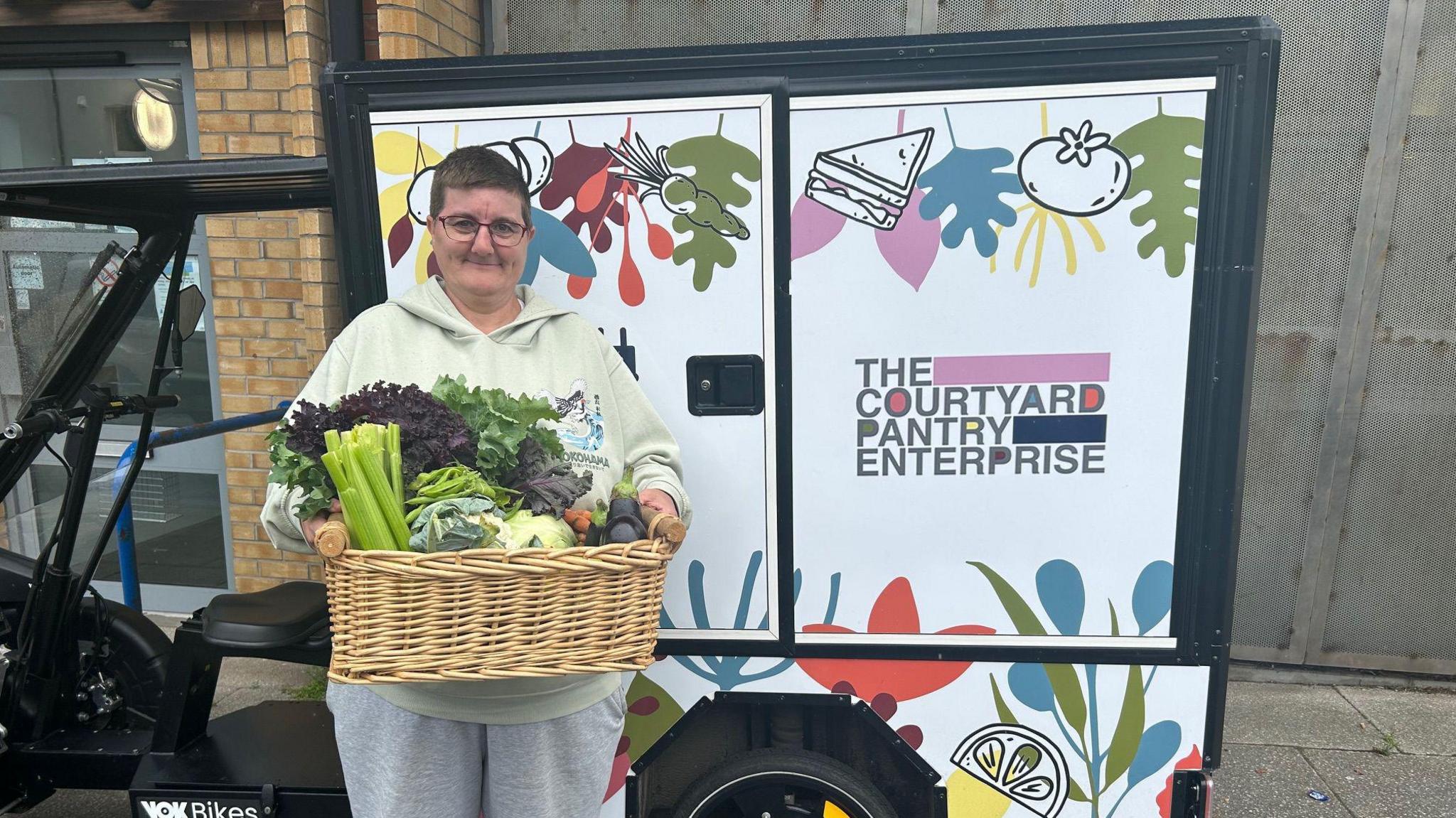 A female volunteer, with glasses and a light green top, holds a basket of vegetables while standing outdoors next to an electric quadricycle, saying THE COURTYARD PANTRY ENTERPRISE on it.