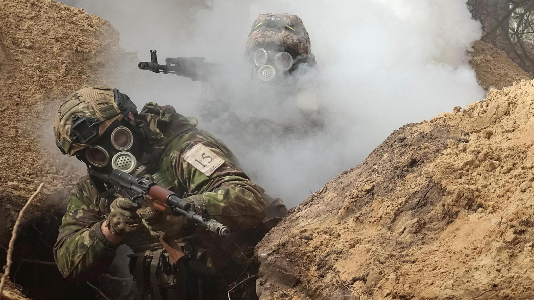 Two Ukrainian soldiers moving through a trench during a chemical weapons training drill. They both have gas masks on and one is partially-obscured by smoke.