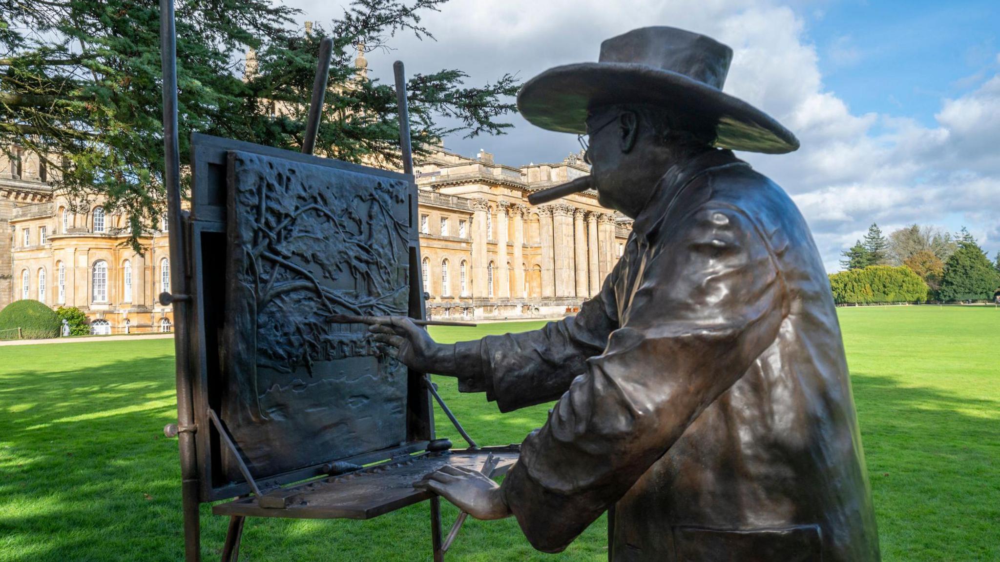 A bronze statue, created by sculptor Paul Rafferty, of Sir Winston painting on the South Lawn of Blenheim Palace. The image is taken from behind the statue with the palace in the background. the sun is shining, casting shadows on the lawn, and the skiy is blue with some clouds to the left.