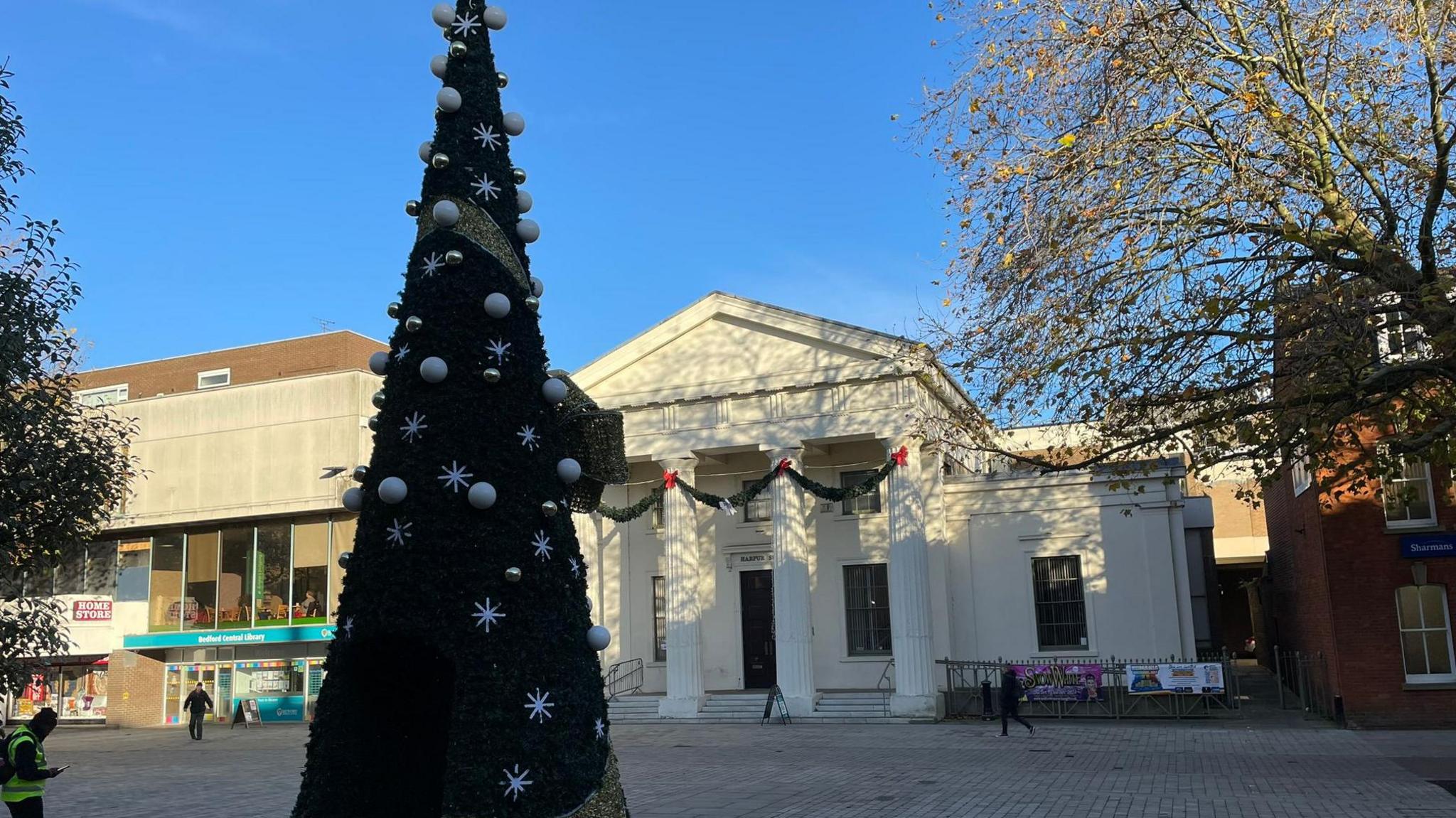 A Christmas tree with white snowflakes and white baubles in a town square. It is in front of a white building which has large columns in front of it. The sky is blue and people are walking in the background.