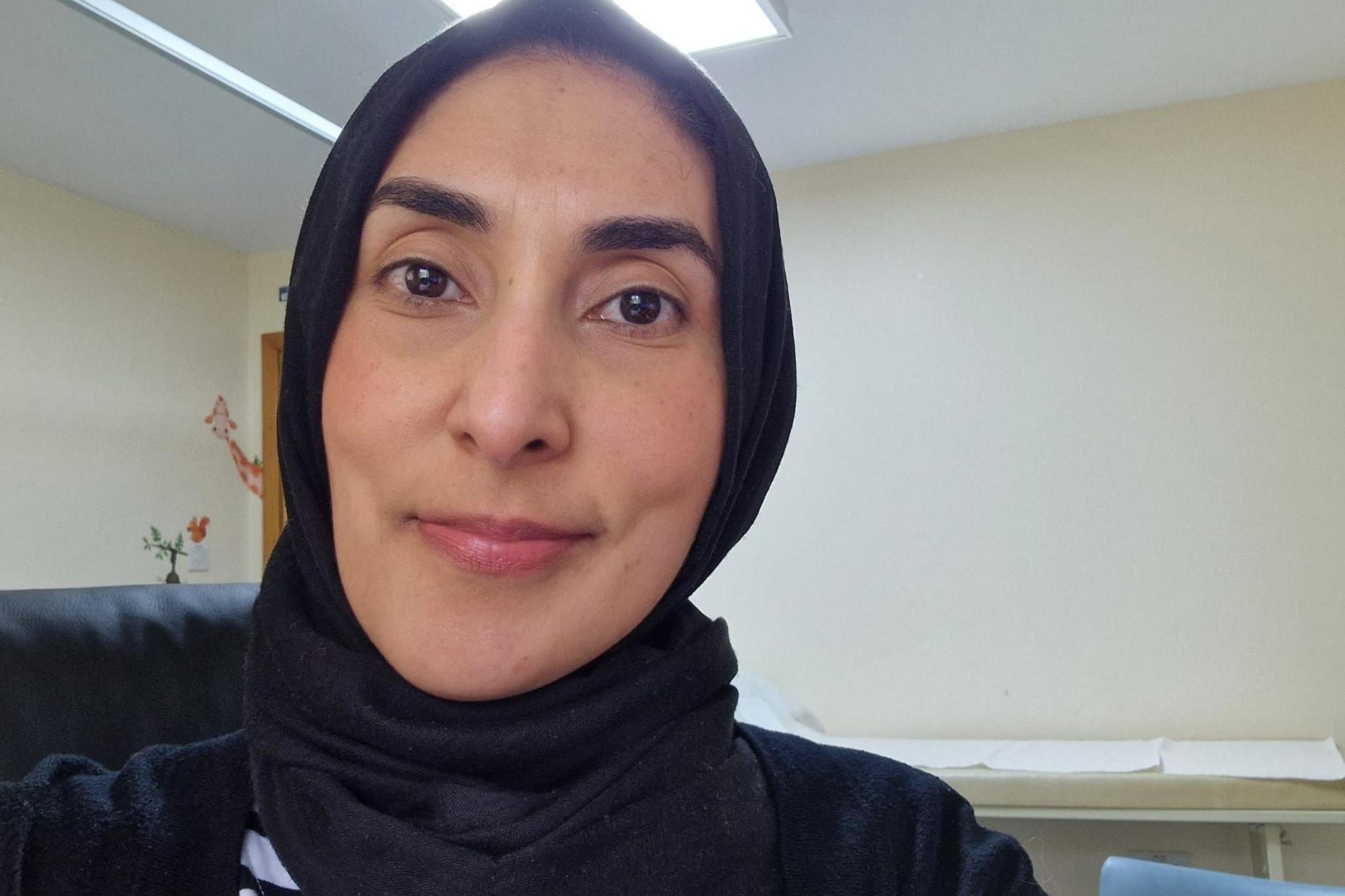 Dr Sahira Dar is smiling at the camera. She is wearing a black hijab and is sitting in a GP office.