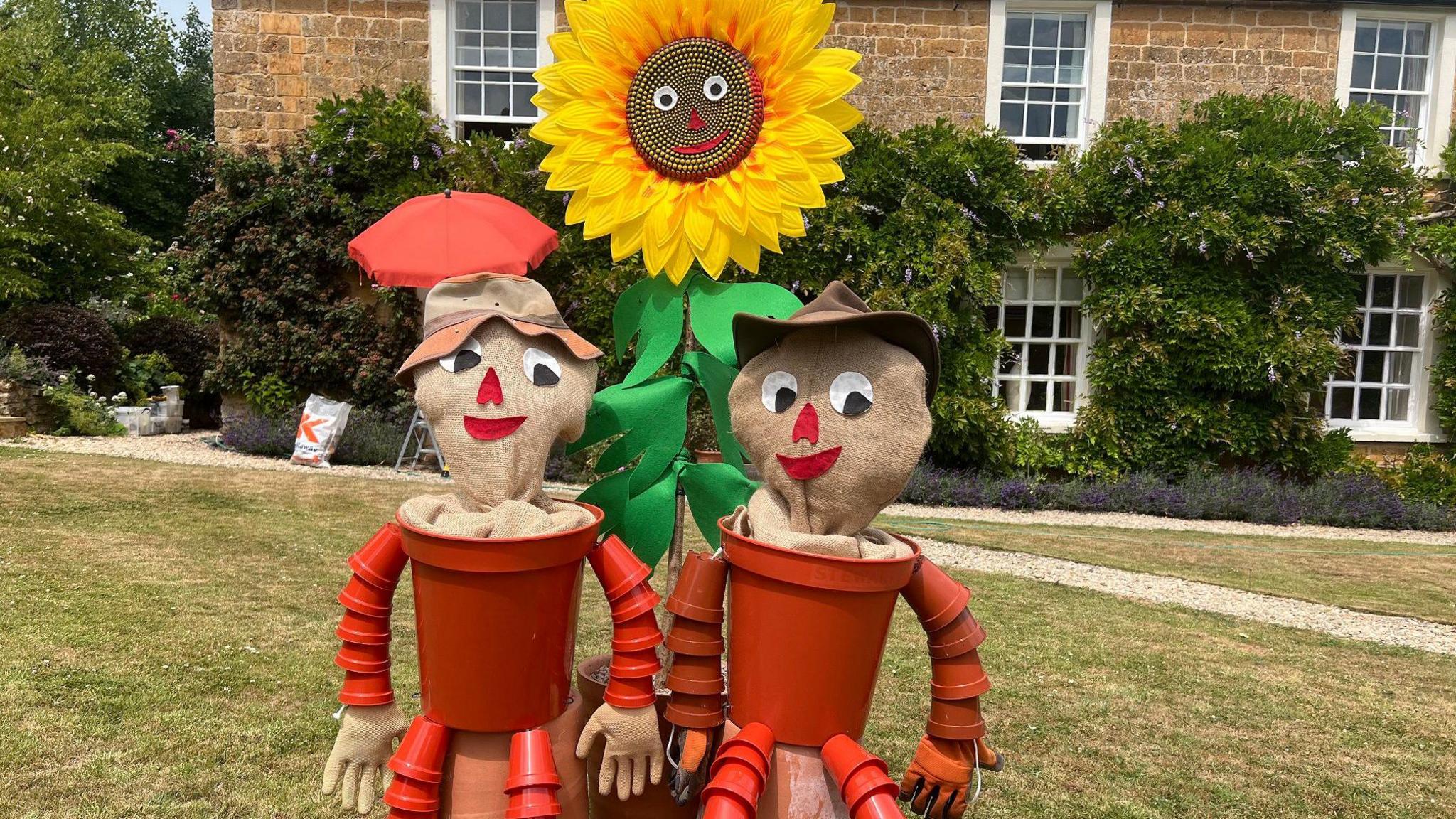 Bill and Ben the Flower Pot Men in scarecrow form sit on a sunflower outside a large country home 