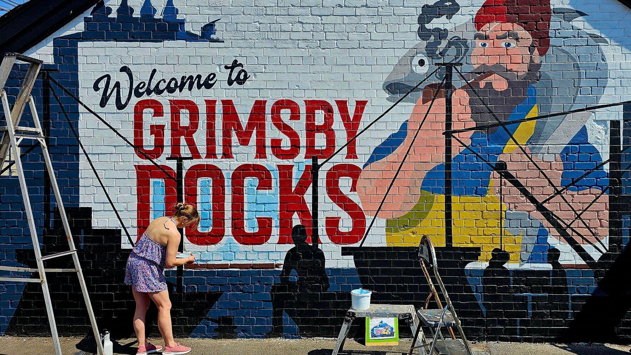 Blonde woman paints details onto a mural for Grimsby Docks, depicting a fisherman holding a fish, with text that reads: Welcome to Grimsby docks