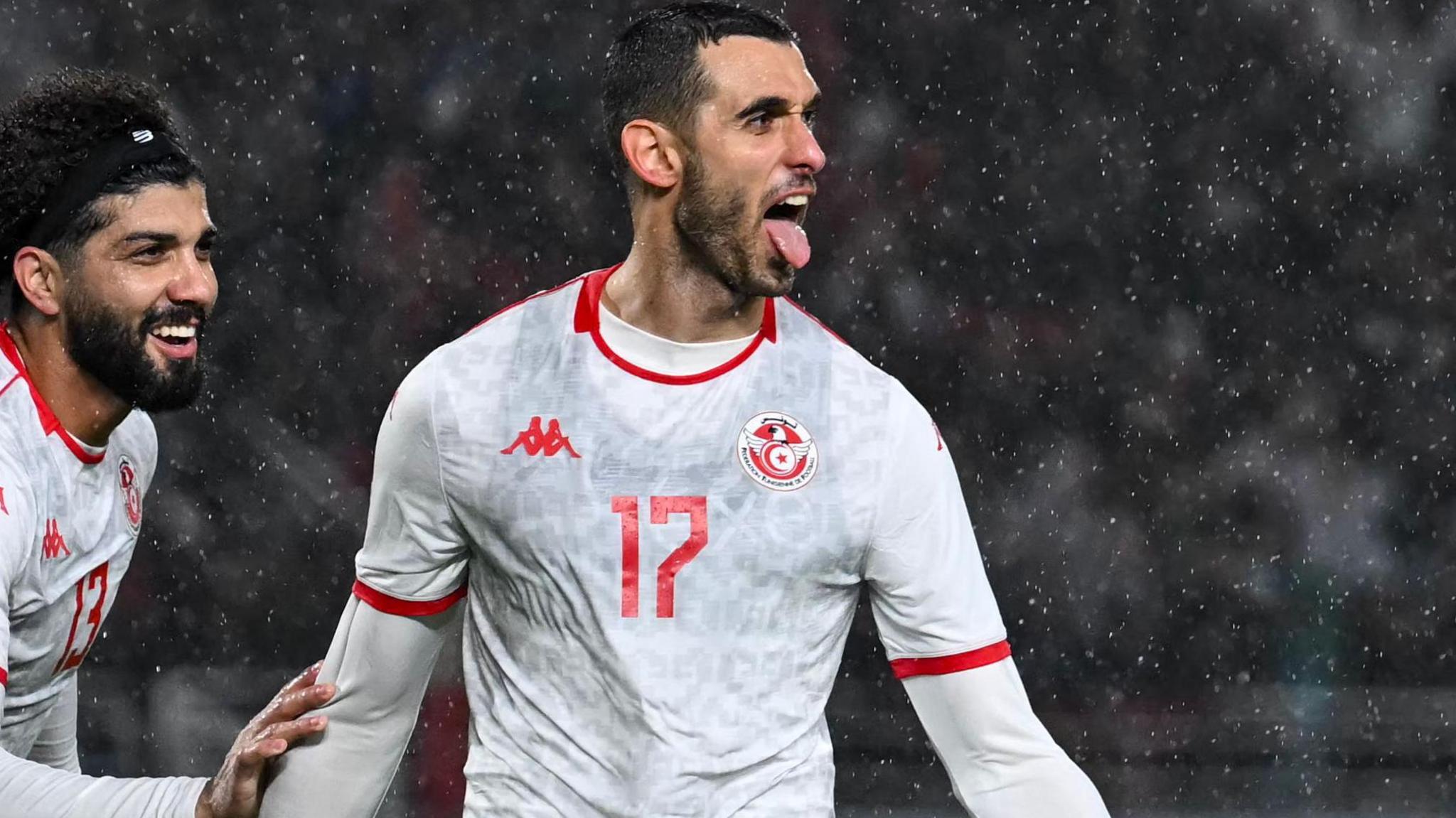 Footballer Ellyes Skhiri celebrates after scoring a goal for Tunisia against Uganda at Afcon 2025. He is visible from waist up in a white away jersey and white under-sleeves, and is extending his tongue. Team-mate Ferjani Sassi, wearing black headband over curly hair, is seen offering his congratulations as he appears from the left-hand side of the image, with large drops of rain seen falling
