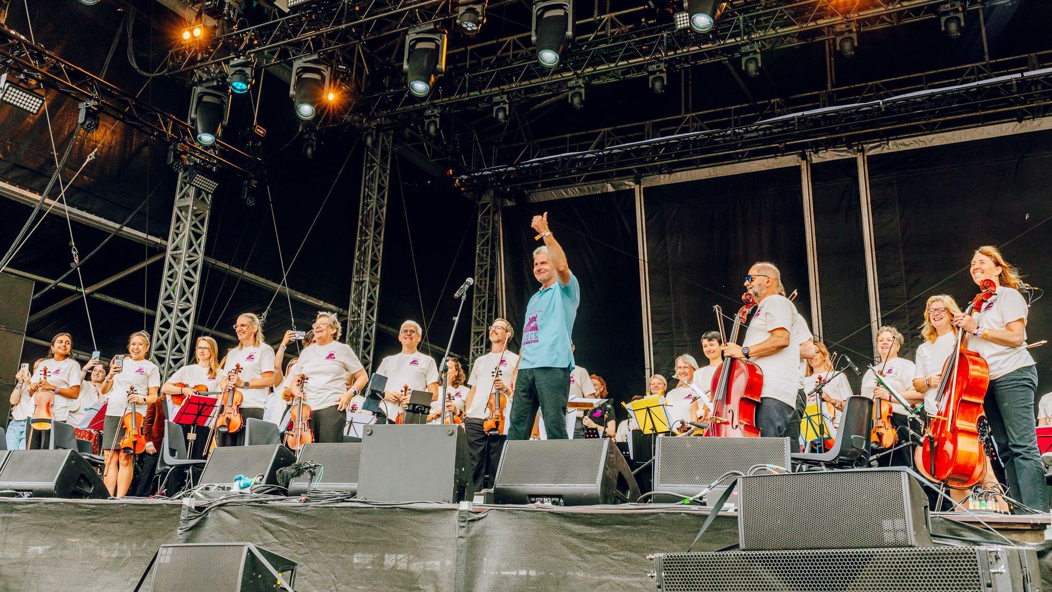 The Oxford Symphony Orchestra on the stage facing the crowd at Truck festival. The members are smiling and holding their instruments.