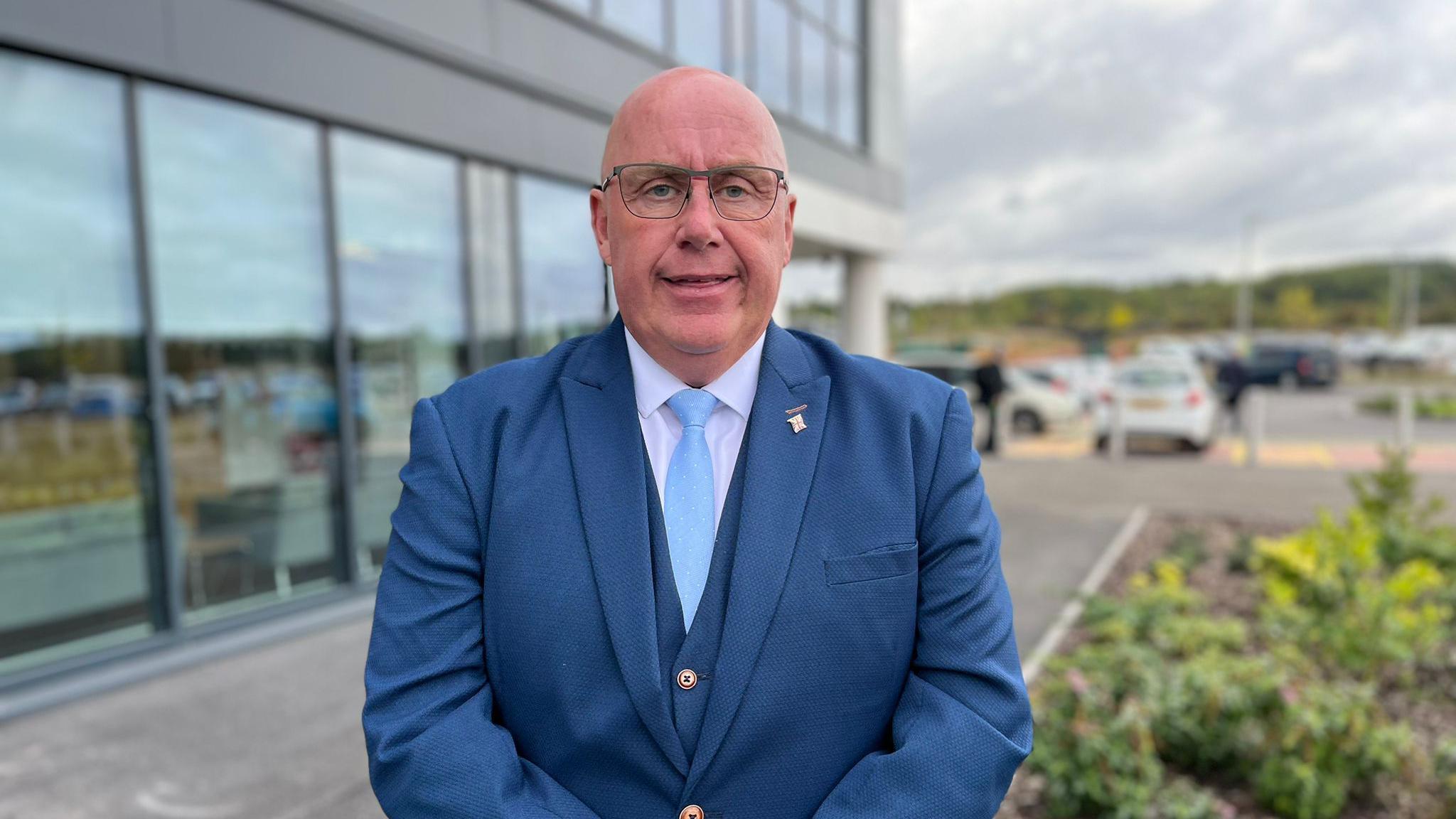 A man in a blue suit and light blue tie. He is bald and is wearing glasses