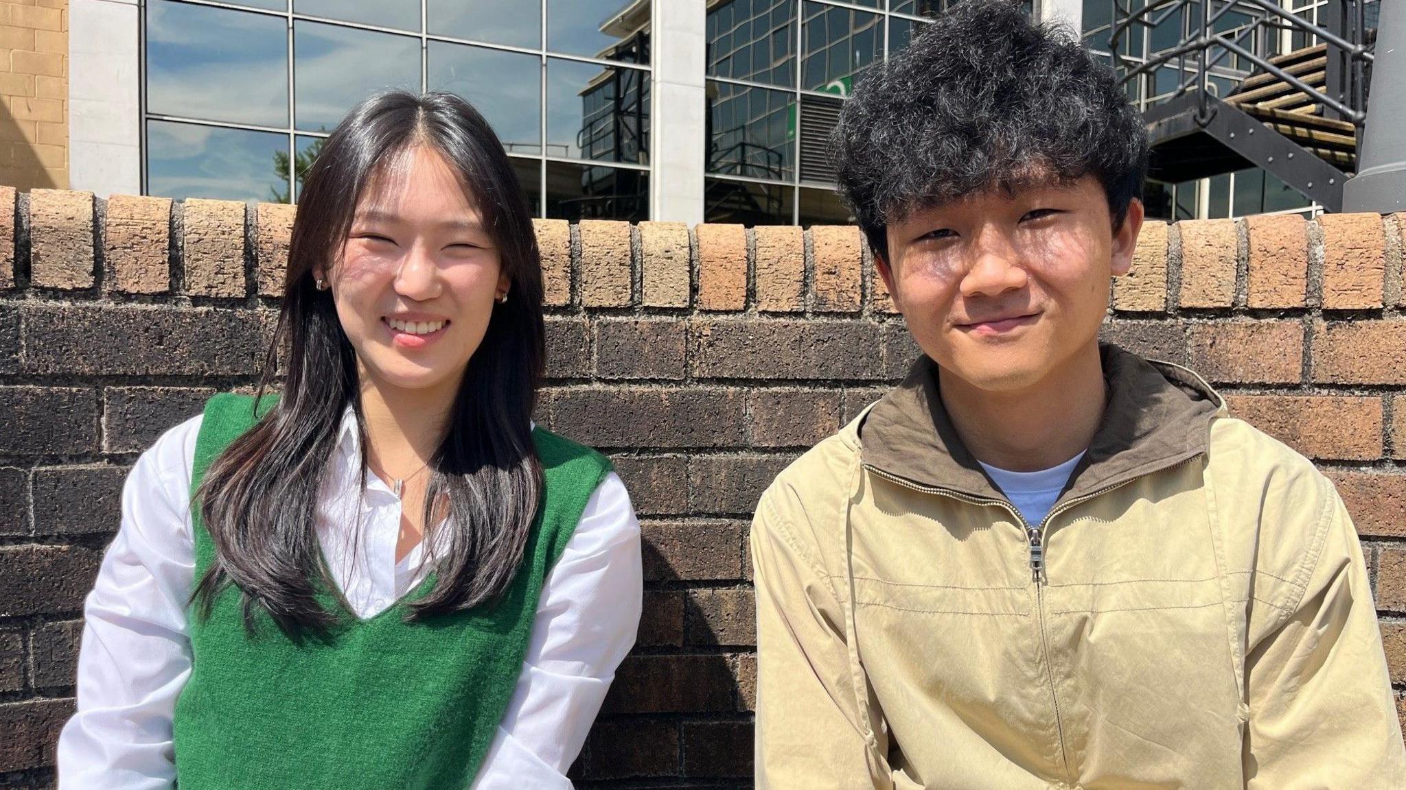 Sunwoo and Sangweon smile at the camera. Sunwoo has long black hair and is wearing a white shirt. Sangweon is wearing a beige vip up jacket and has short black hair. They are seated with a brick wall behind them.