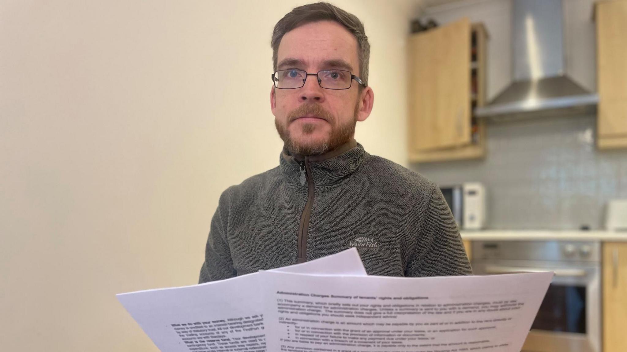 A man in a grey top and glasses holds up a couple of pieces of paper; a kitchen is pictured in the background.