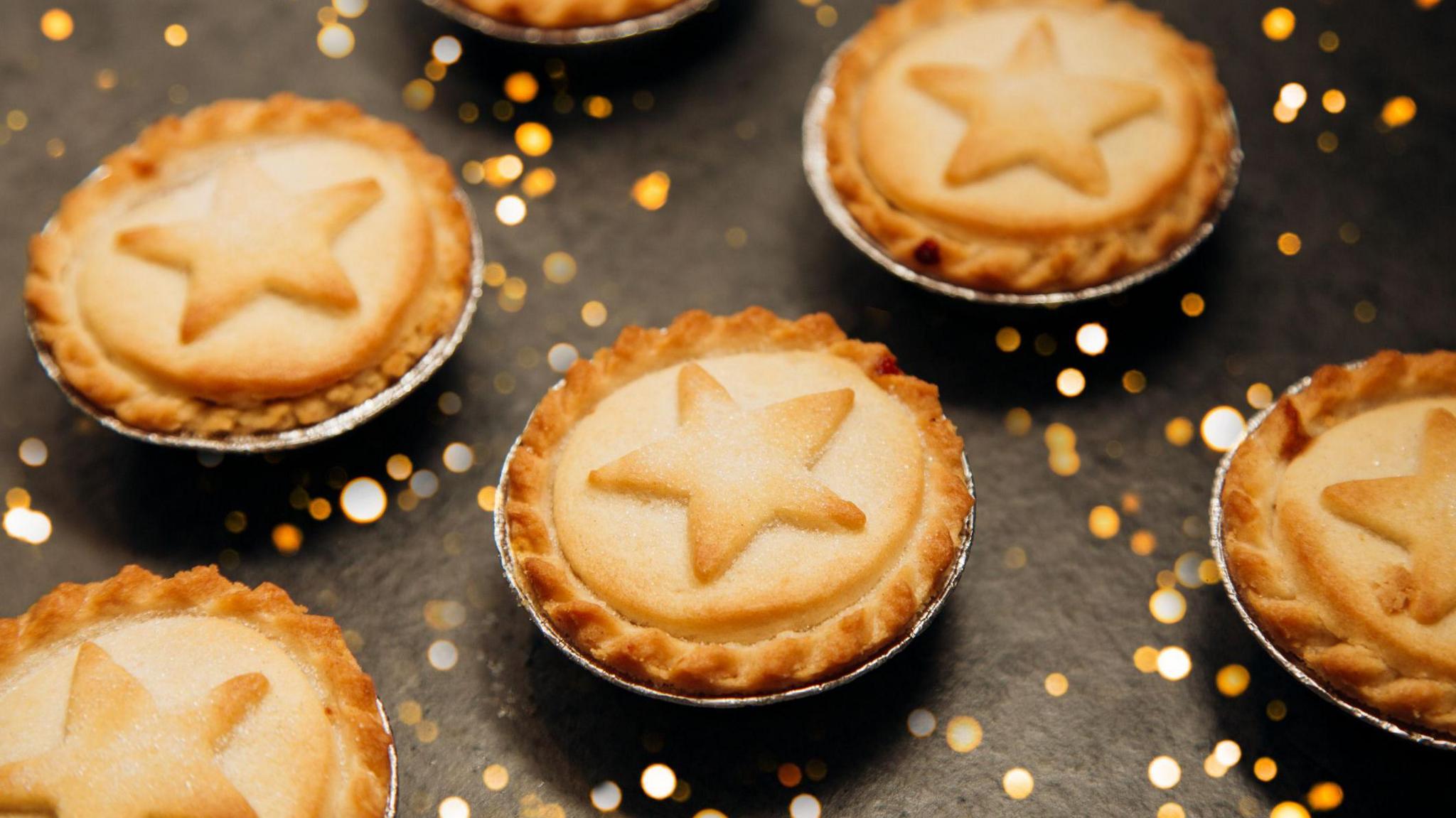 Shortcrust mince pies are laid out evenly on a black surface with sparkly confetti on it. They are in small, round, silver trays. They have a twisted rim of pastry and pastry stars in the middle and sprinkled with sugar. 
