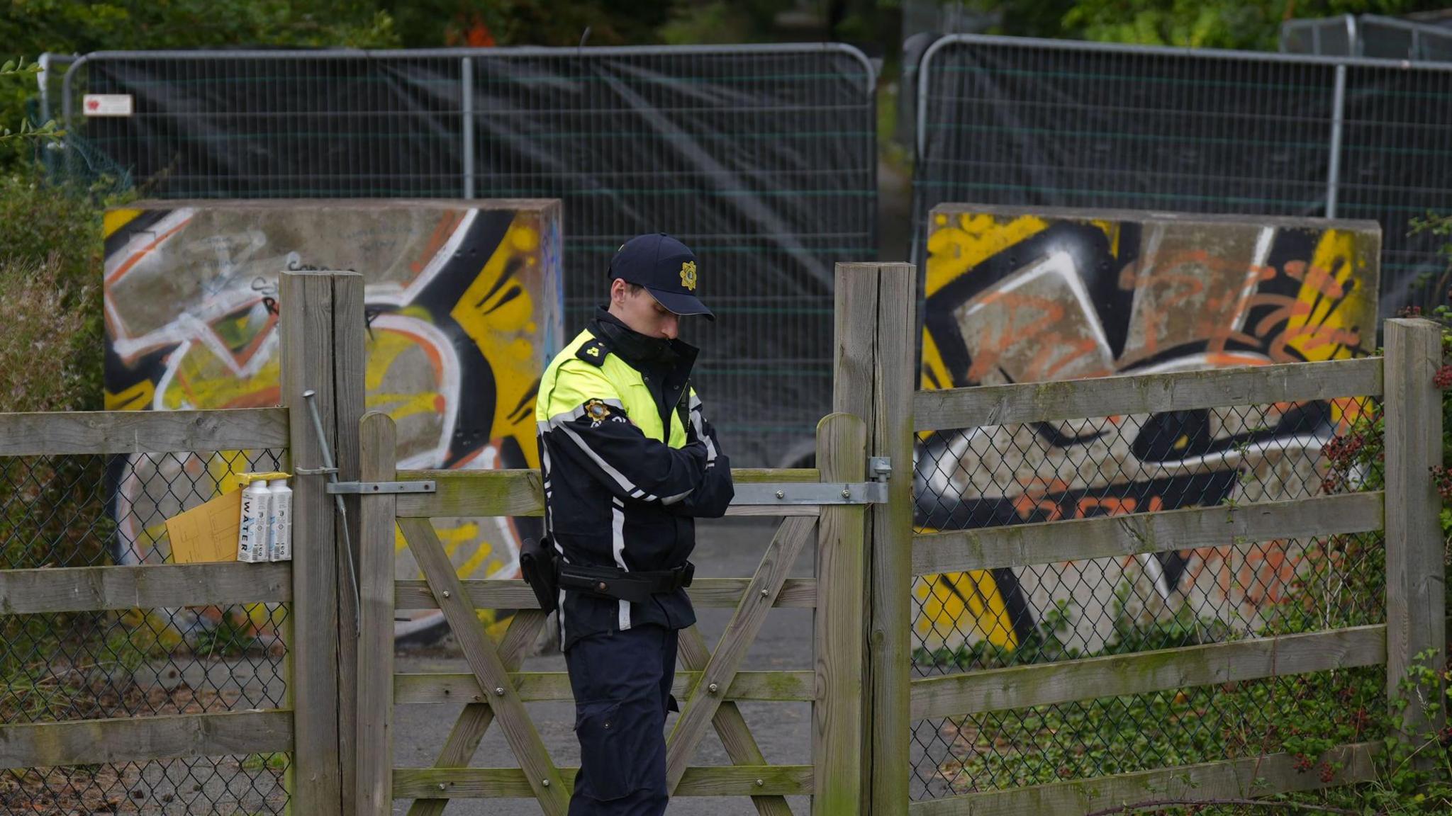 A man wears a black and hi-viz yellow jacket with black trousers. He wears a navy baseball cap. Behind him is a fence with a black sheet on it. protecting an area of green woodland.