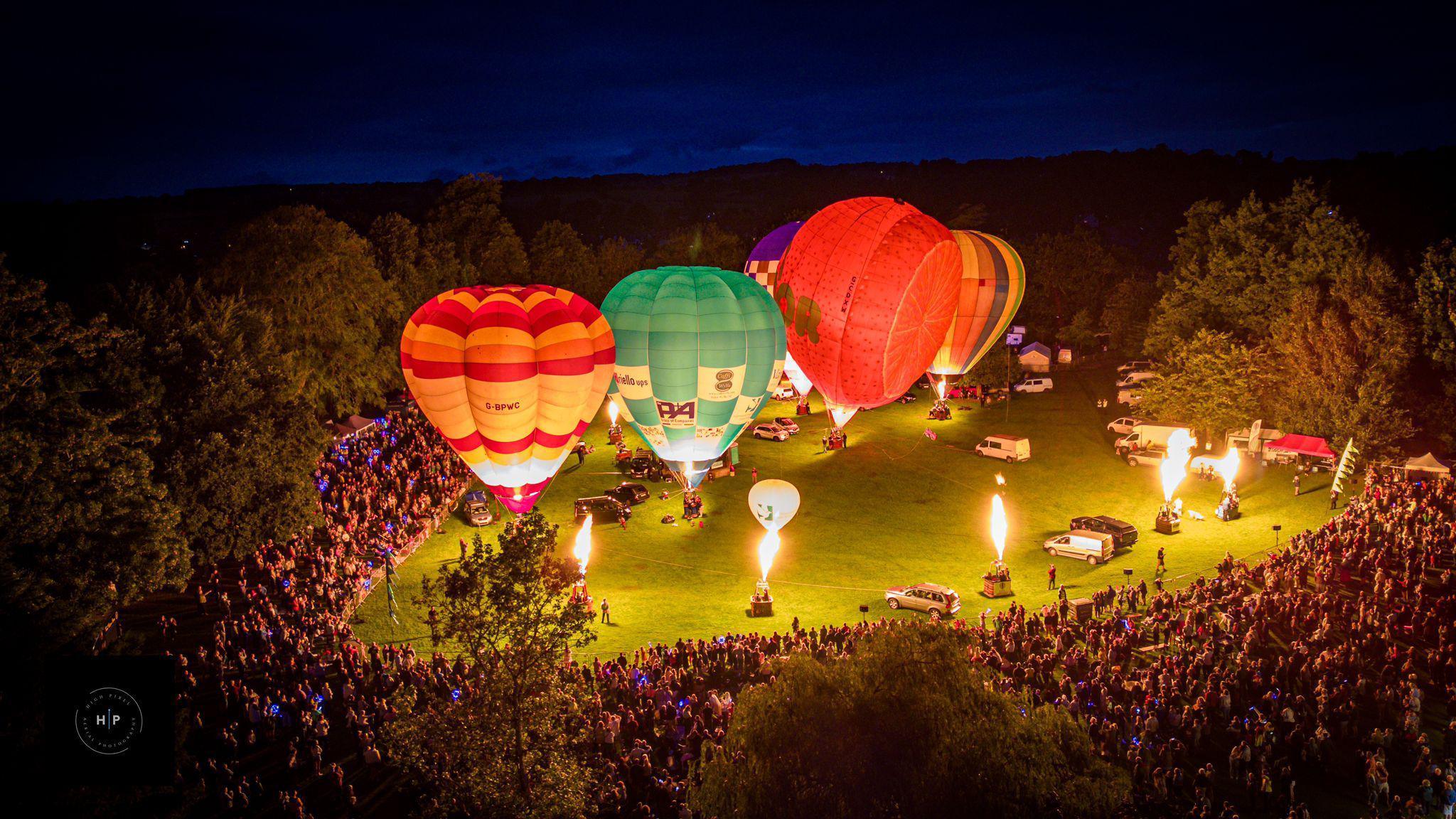 An aerial image taken from above the crowds watching at least five hot air balloons inflate at night. The balloons are of various sizes and shapes and there are baskets around the balloons which are shooting fire into the sky. 