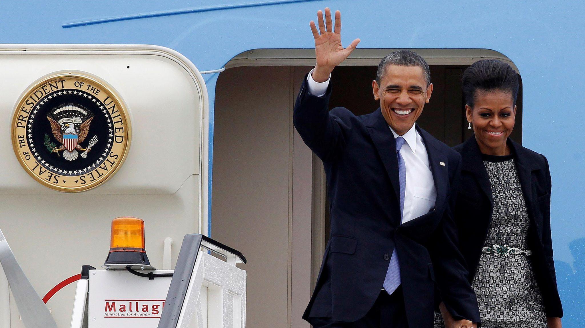 US President Barack Obama and first lady Michelle Obama arrive aboard Air Force One at Dublin airport. Barack is wearing a dark suit, white shift and blue tie and he is waving. Michelle is wearing a black and white patterned dress and a black blazer.