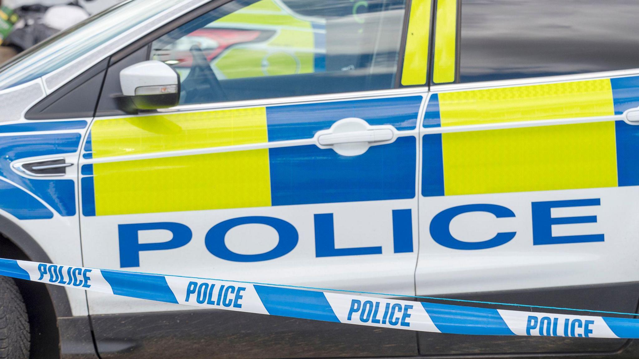 The side view of a police car with blue and yellow markings. A blue and white police cordon crosses in front of it.