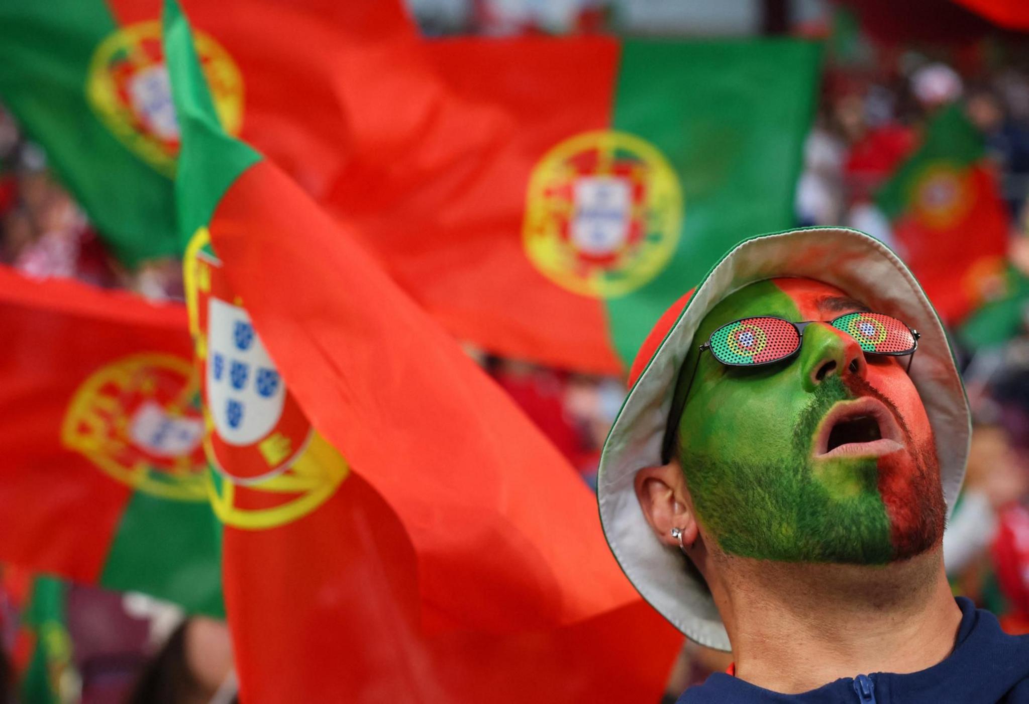 A Portuguese fan wearing full face paint and sunglasses looks up to the sky in front of a sea of Portuguese flags
