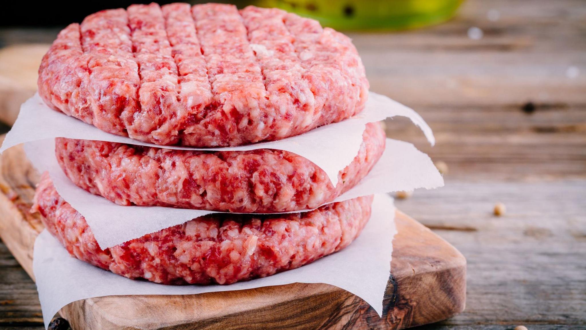 Three beef burgers separated by paper on top of a wooden chopping board and an wooden table