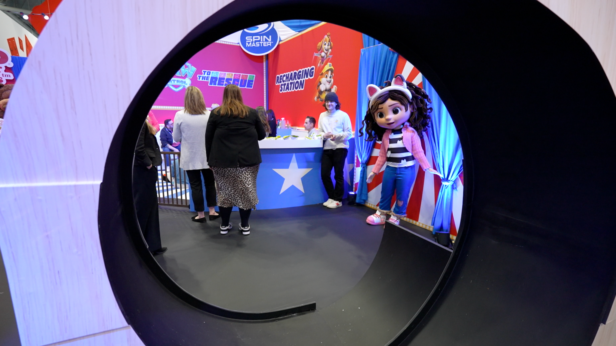 A display at the toy fair with people clustered around the booth from the back and a giant doll with cat ears