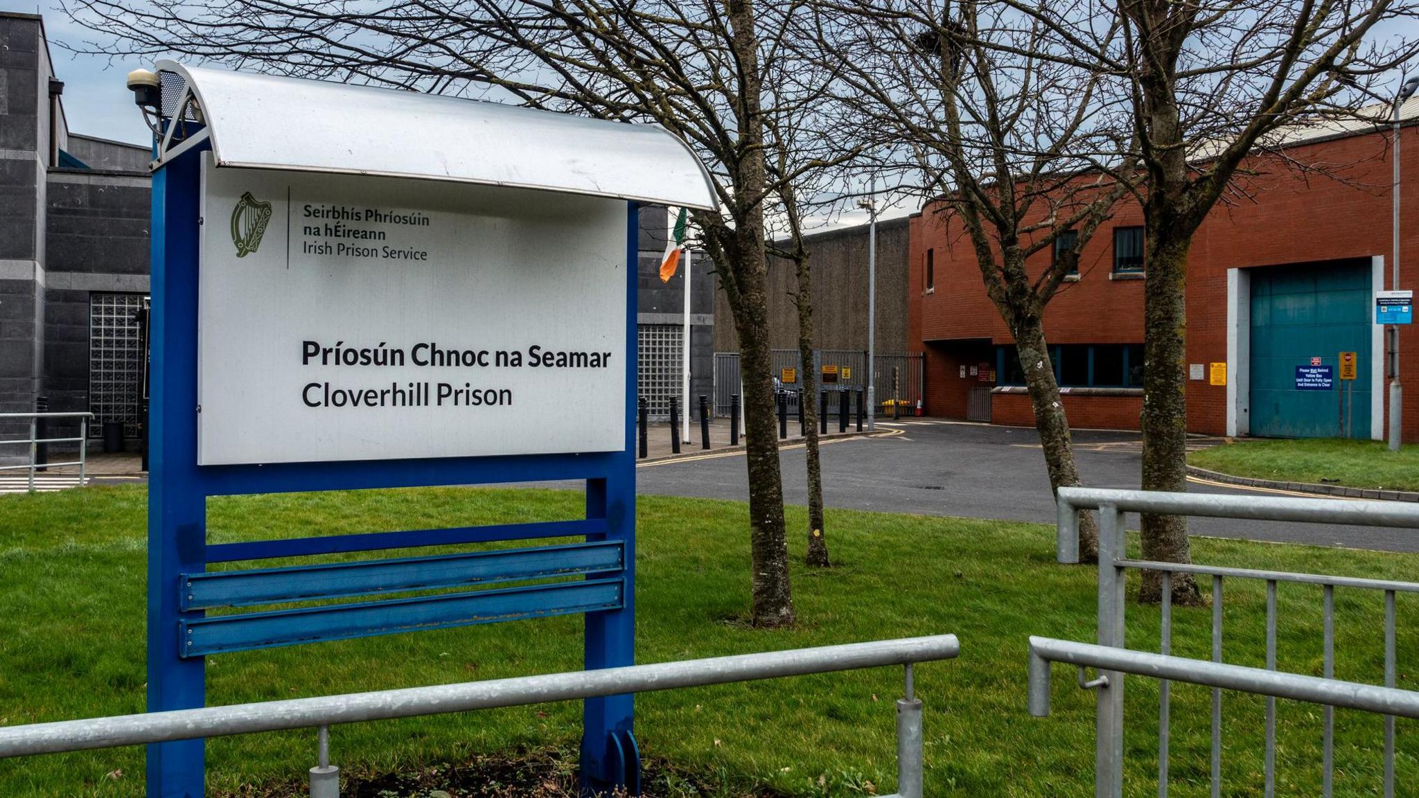 A blue and white sign with "Cloverhill Prison" written on it surrounded by grass, metal barriers and trees. In the background is a brown and dark grey building.