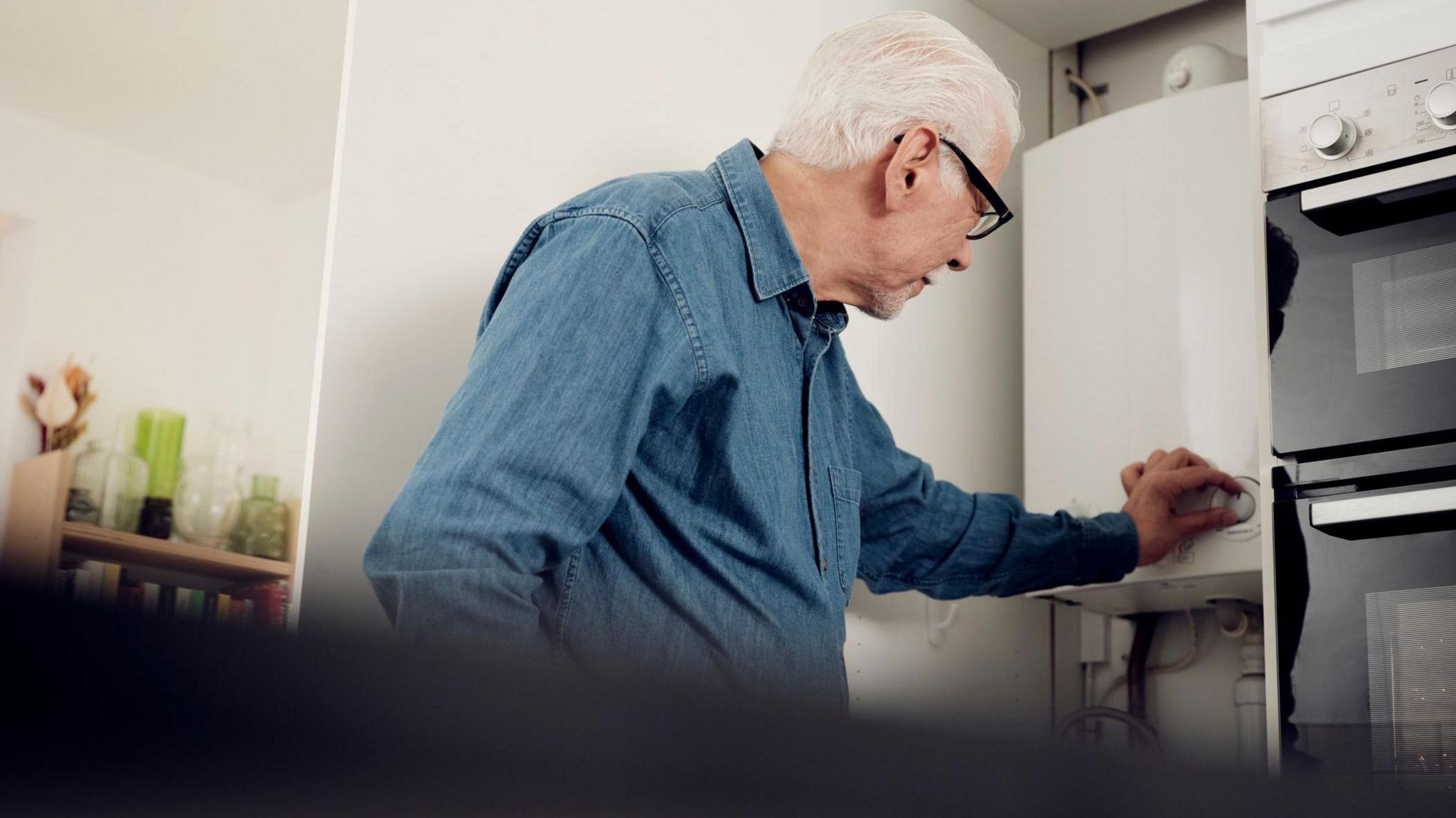 Older man has his hand on the controls of a boiler in a kitchen.