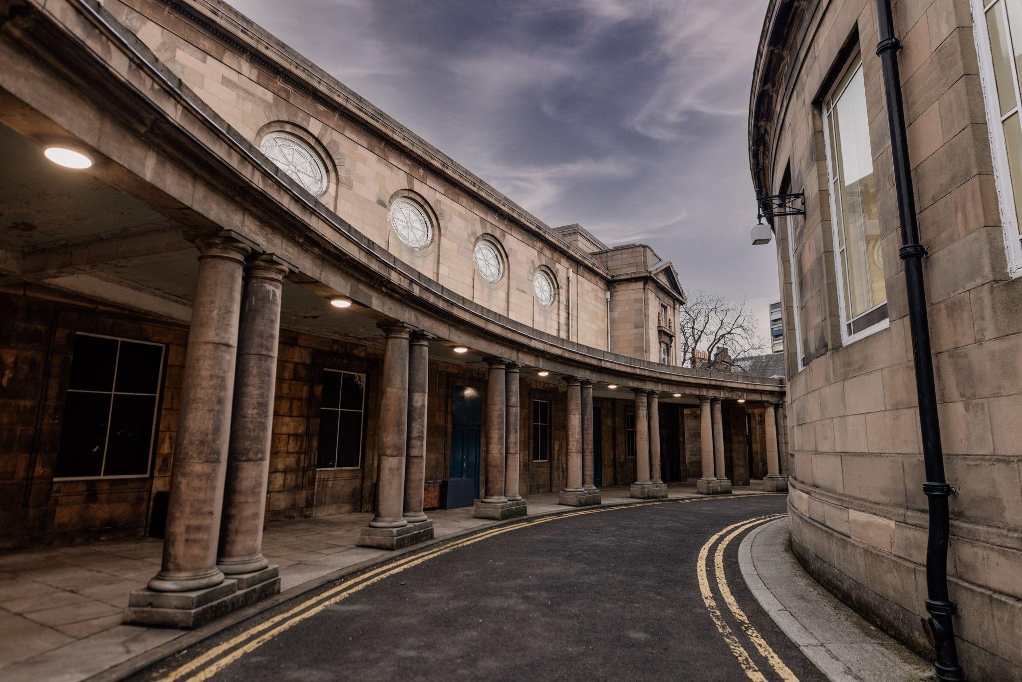 A curved road outside a sandstone building with a columned walkway an round windows above it