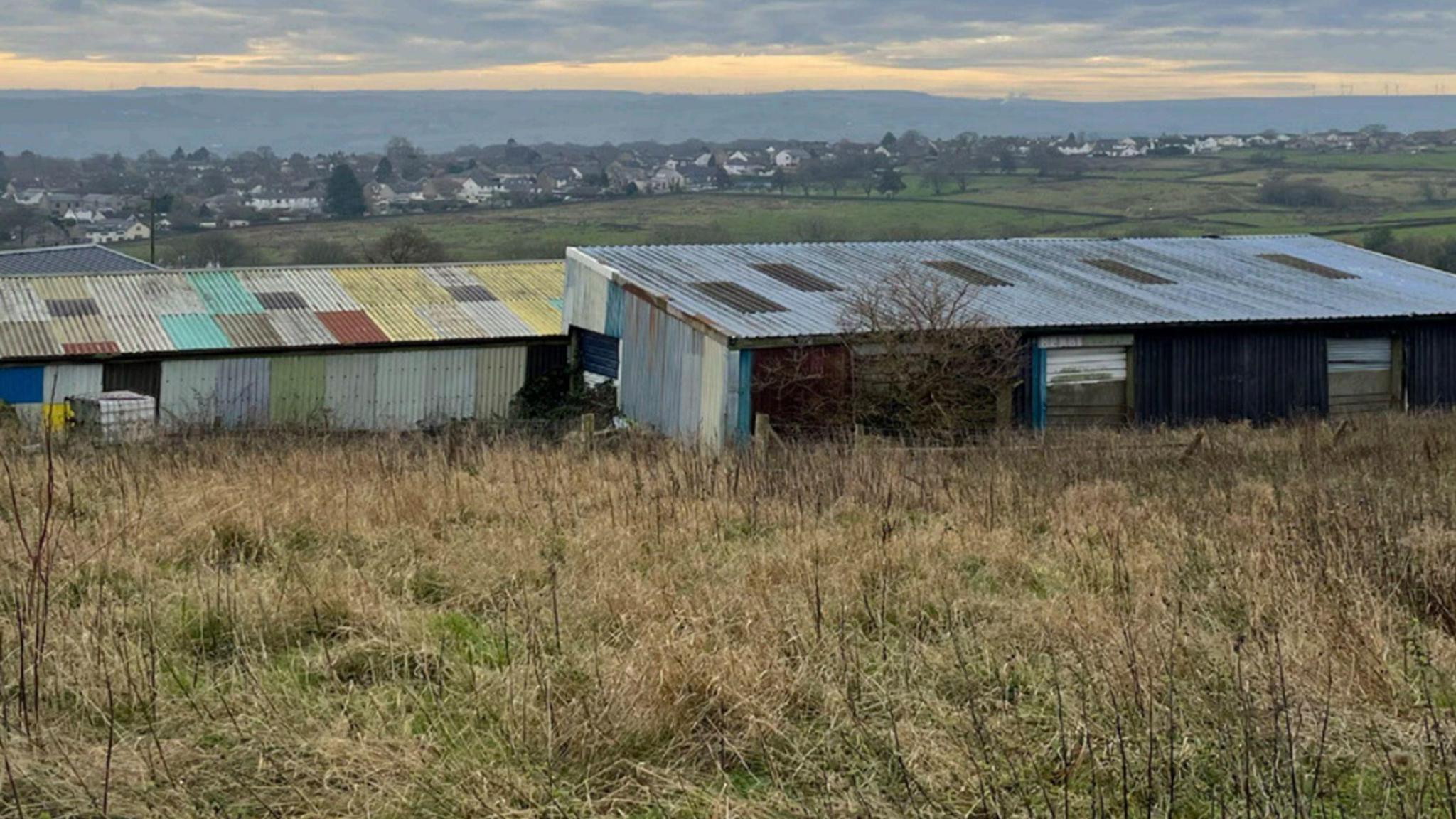 Corrugated iron farm buildings on empty green land overlooking hills with houses in the distance
