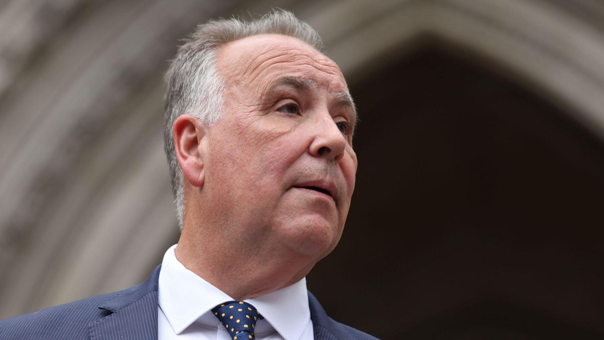 Chris Whitbread is dressed in a formal dark blue suit paired with a white shirt and a navy tie featuring a pattern of small yellow dots. He is standing in front of point archways belonging to the High Court in London.