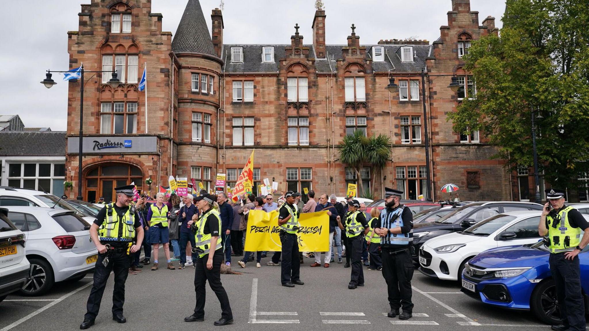 A group of protestors holding signs which say things like "Perth against racism" stand by the entrance to the Radisson Blu hotel. The entrance leads on to a car park with parked cars. Police stand closer to the camera, in front of the protestors.