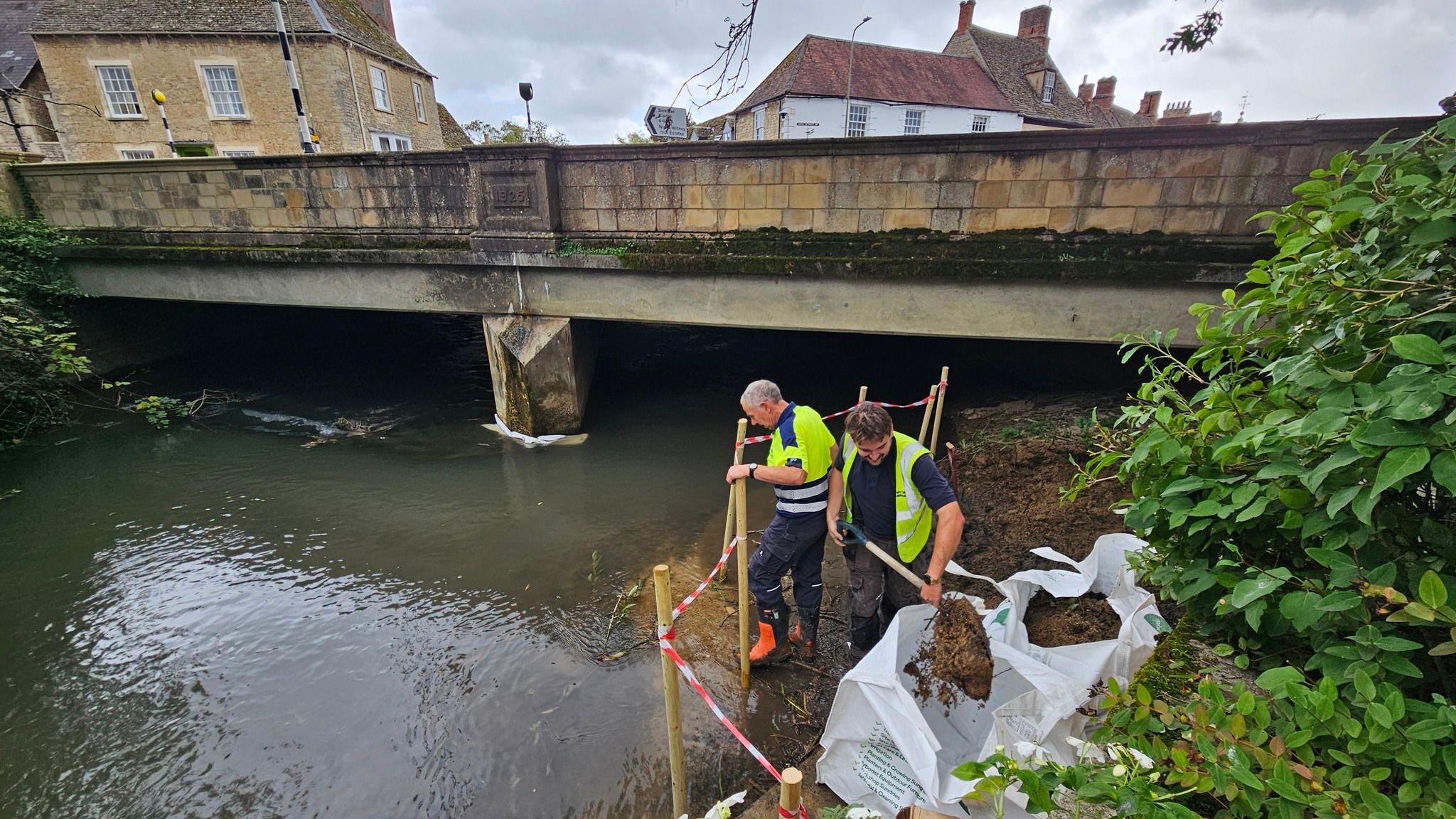 Two workers desilting by the river at Witney Bridge Street, It is a cloudy day.