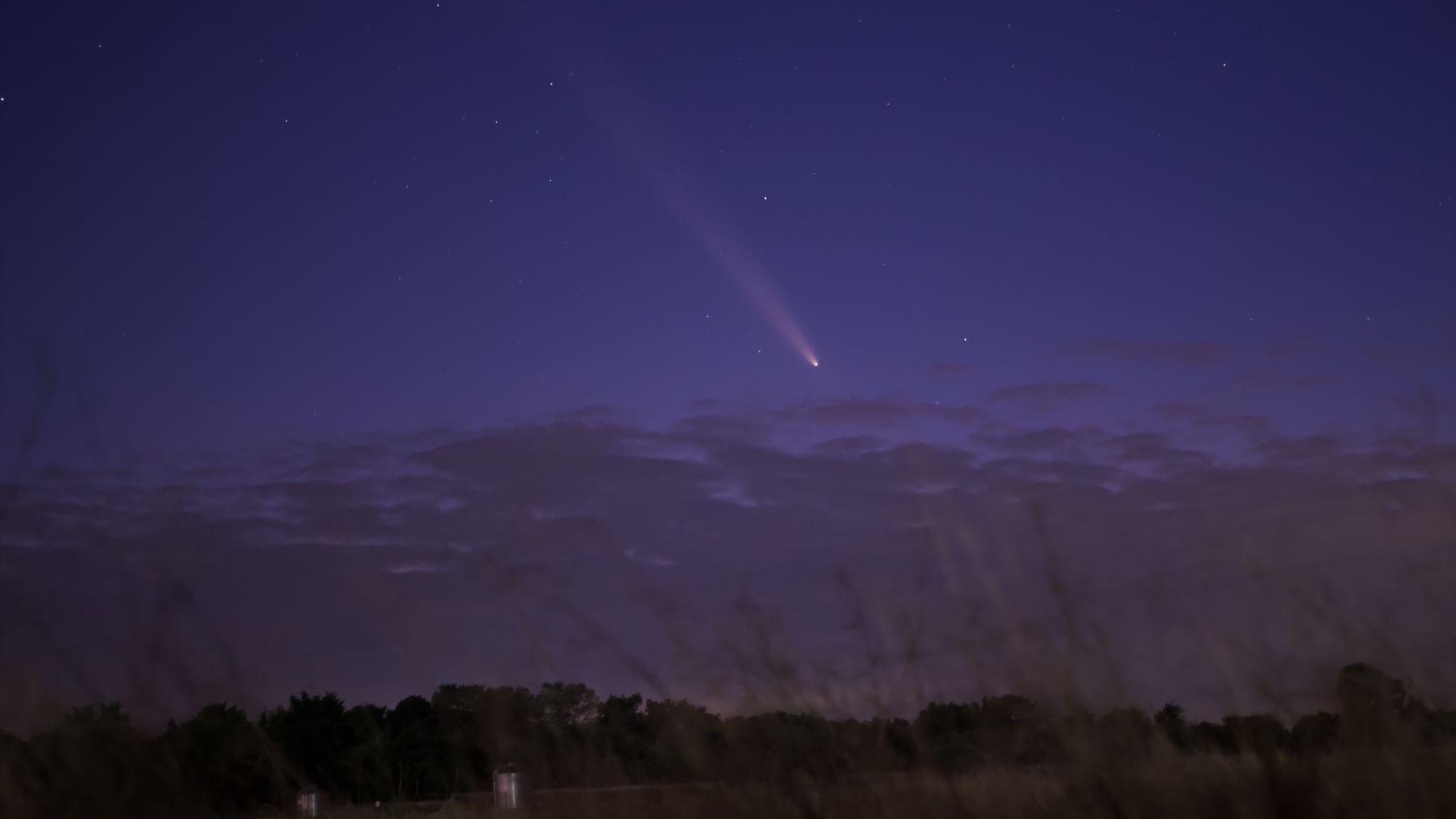 Clouds can be seen on the horizon as the comet travels through the night sky. There are many other stars in the sky.