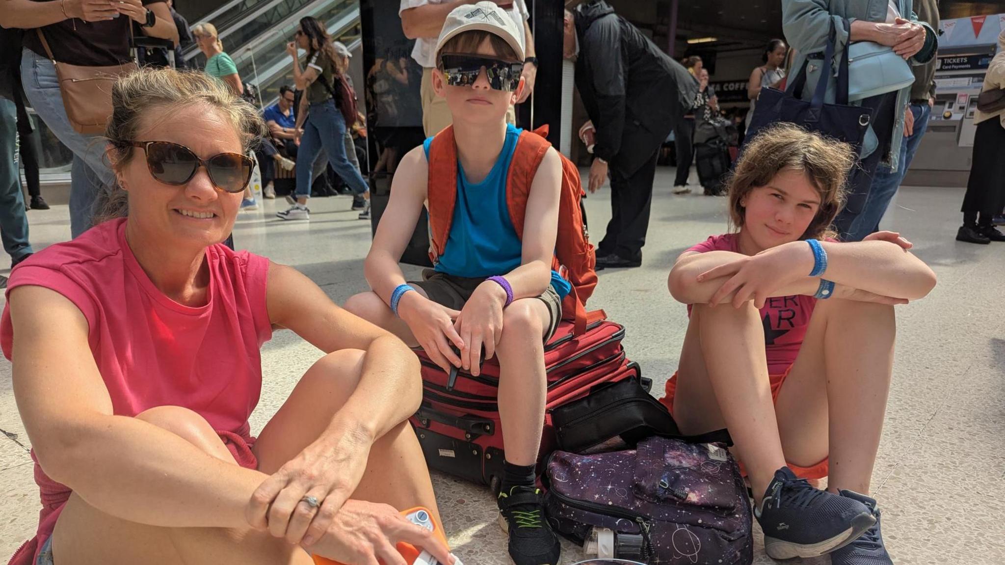 A woman in a pink top and sunglasses sits on the station concourse with two children, a girl and boy ages about 10-14