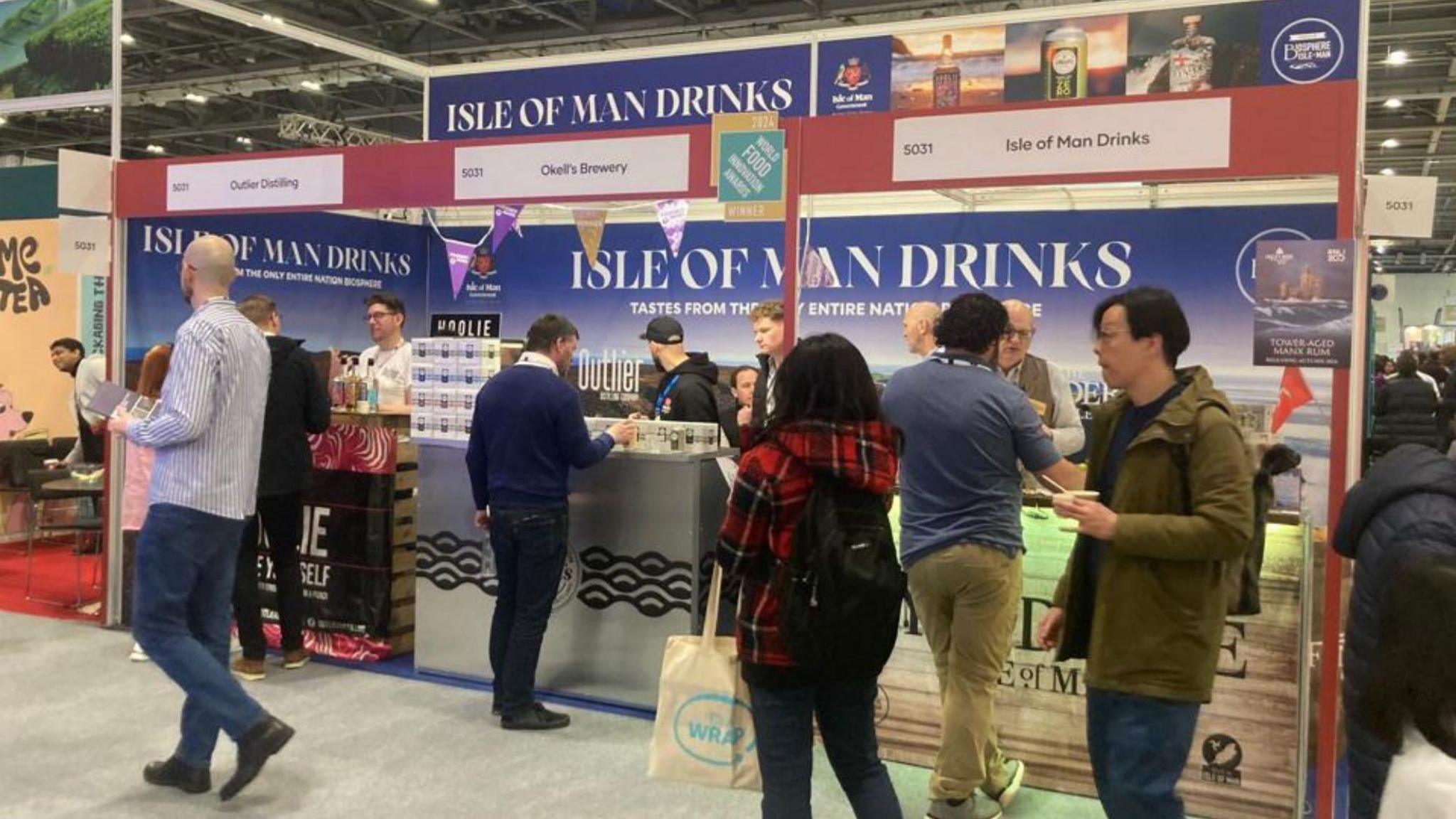 Members of the public milling about at a stand that says Isle of Man Drinks, with some men serving them.