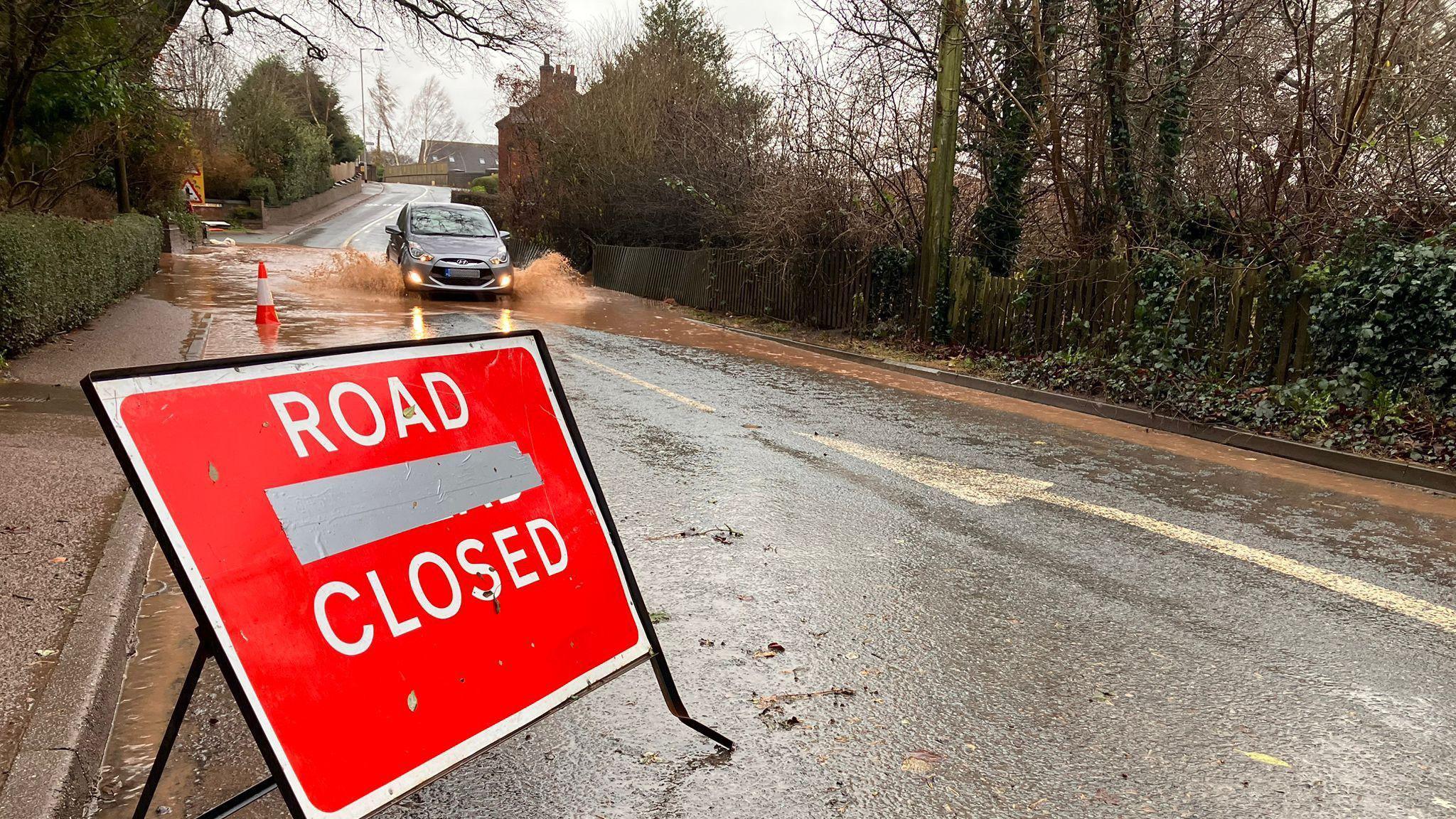 A red "road closed" sign on the side of Bar Hill which is flooded with a silver car driving through the brown water