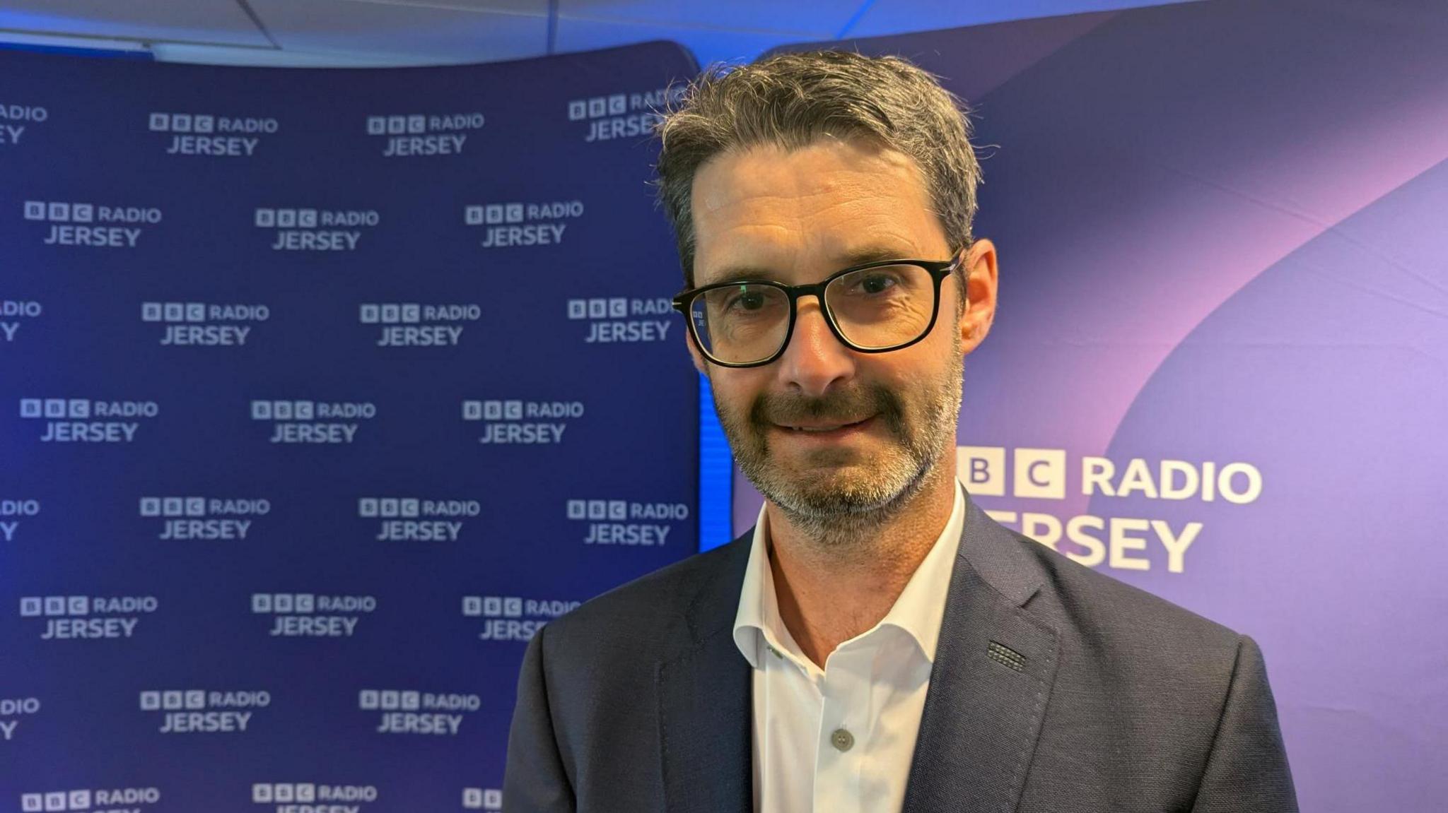 A man in a suit standing in front of a backdrop that repeatedly displays the text "BBC RADIO JERSEY."