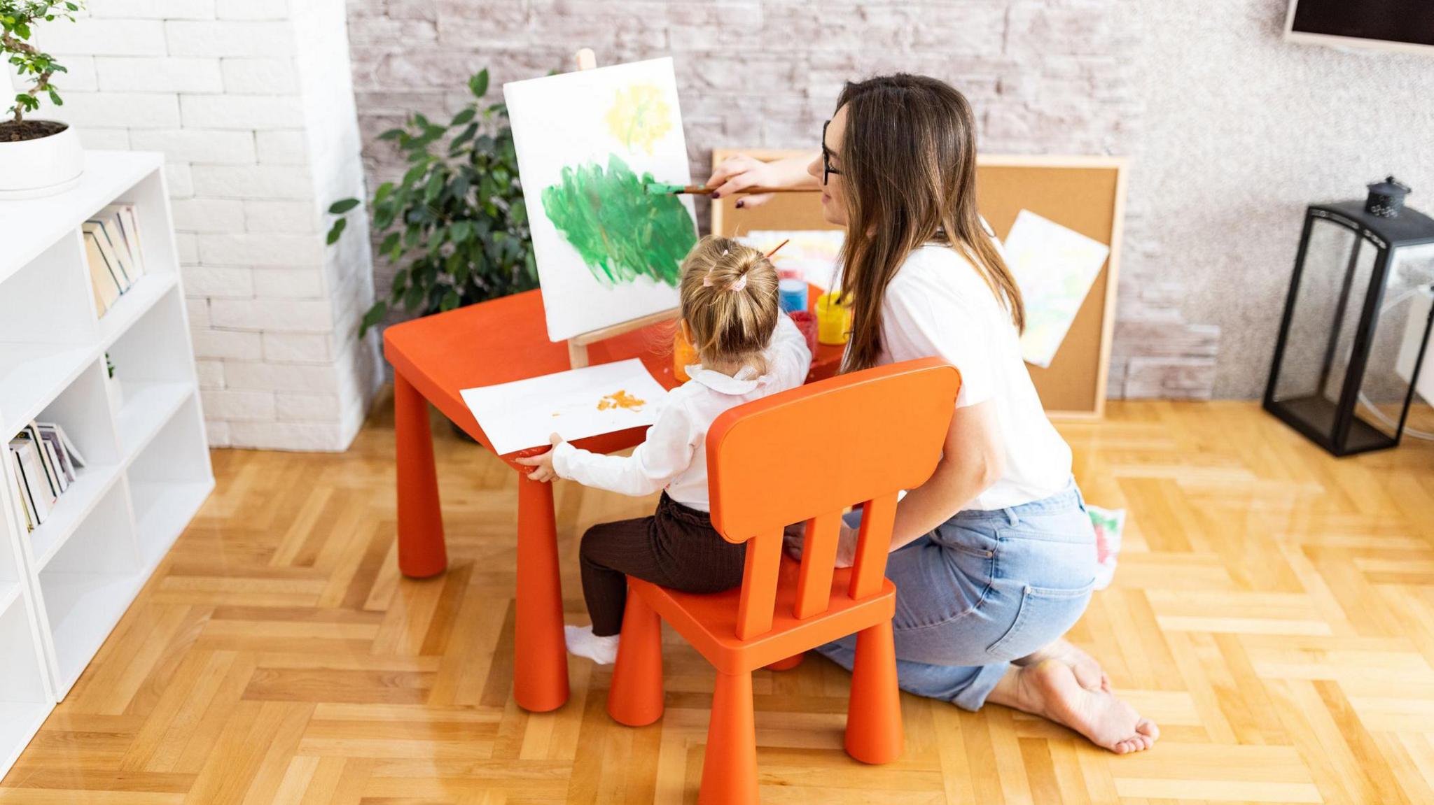 A woman kneels on the floor next to a small child who is painting at a table